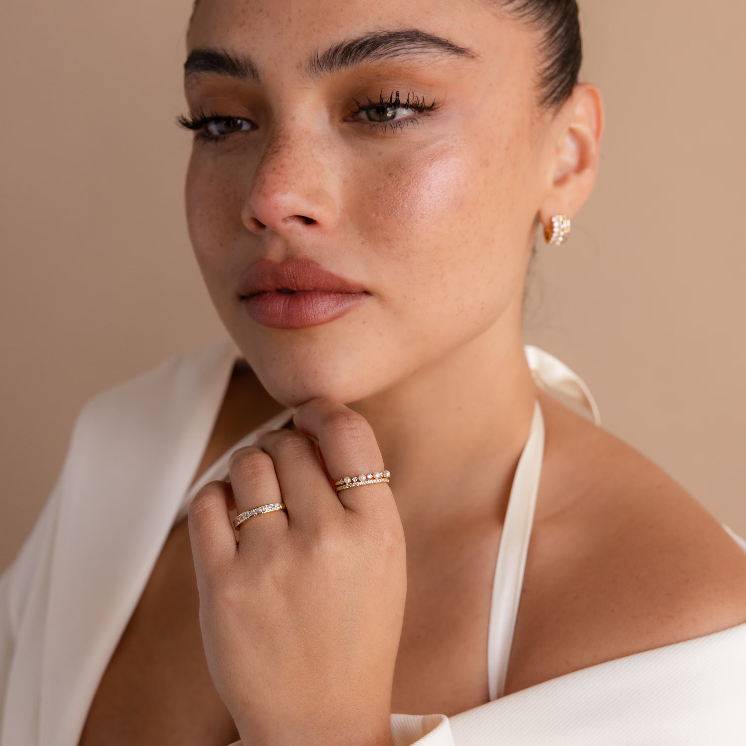 Woman with dewy skin gazing confidently, wearing a white halter top and the Pave Pearl Stacking Ring Set, along with gold gemstone rings and an earring.