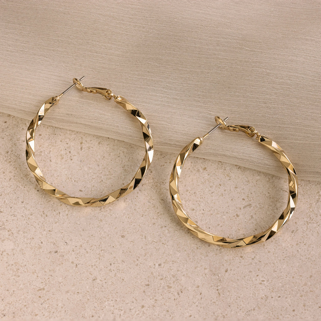 Large Twisted Hoops sit on a beige stone with light fabric draped above—an eye-catching summer accessory that adds effortless elegance.