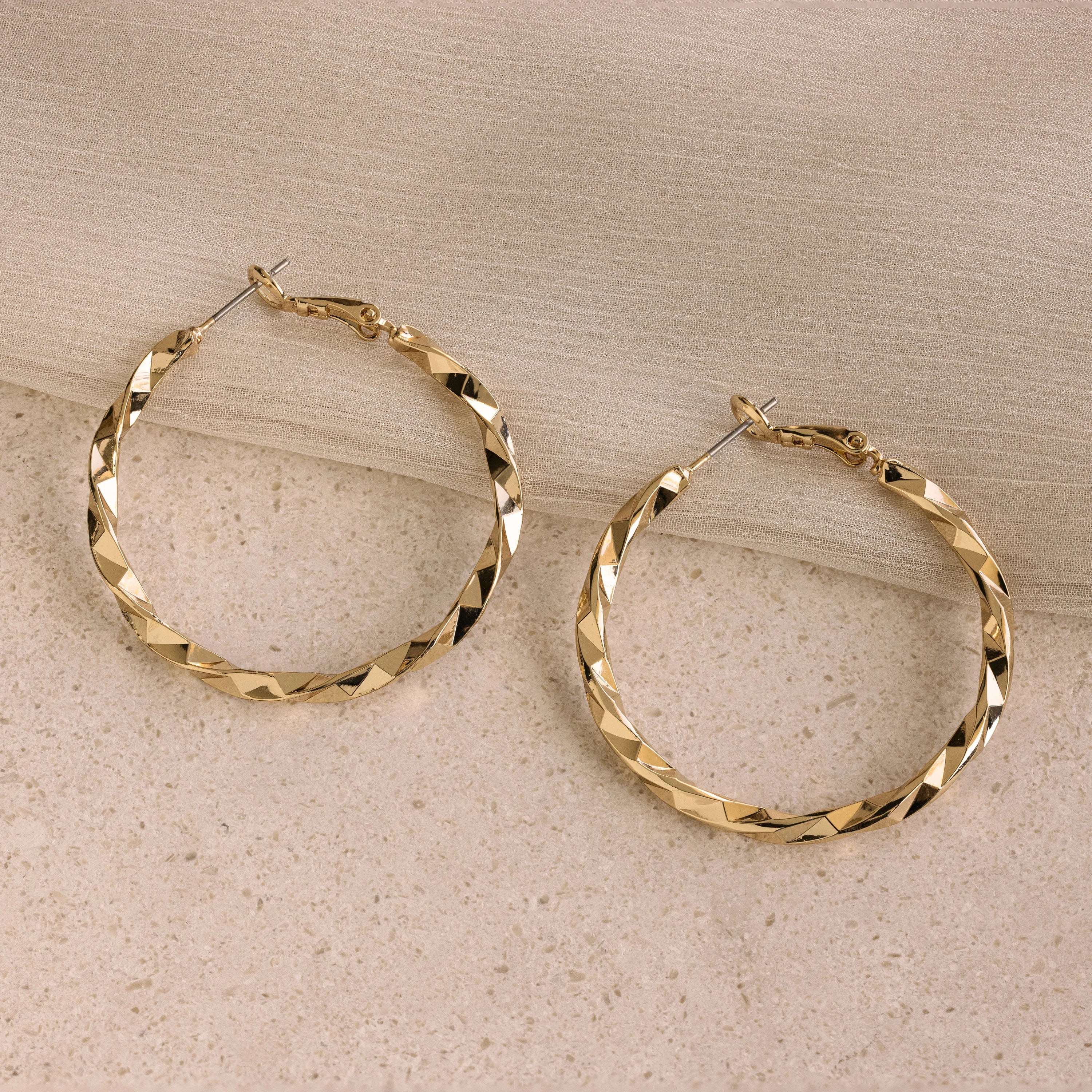 Large Twisted Hoops sit on a beige stone with light fabric draped above—an eye-catching summer accessory that adds effortless elegance.