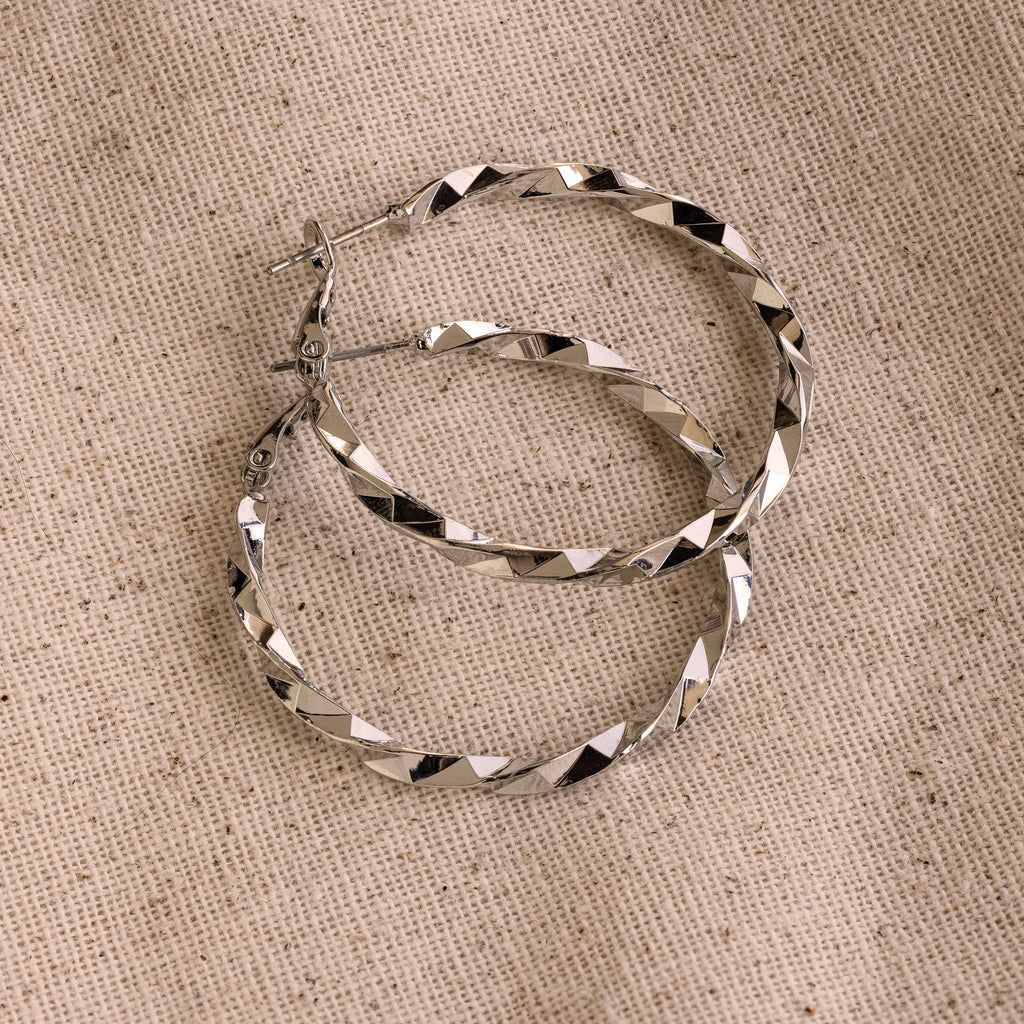 The Large Twisted Hoops, in silver, rest on a beige woven fabric background—an ideal choice for summer style.