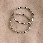 The Large Twisted Hoops, in silver, rest on a beige woven fabric background—an ideal choice for summer style.