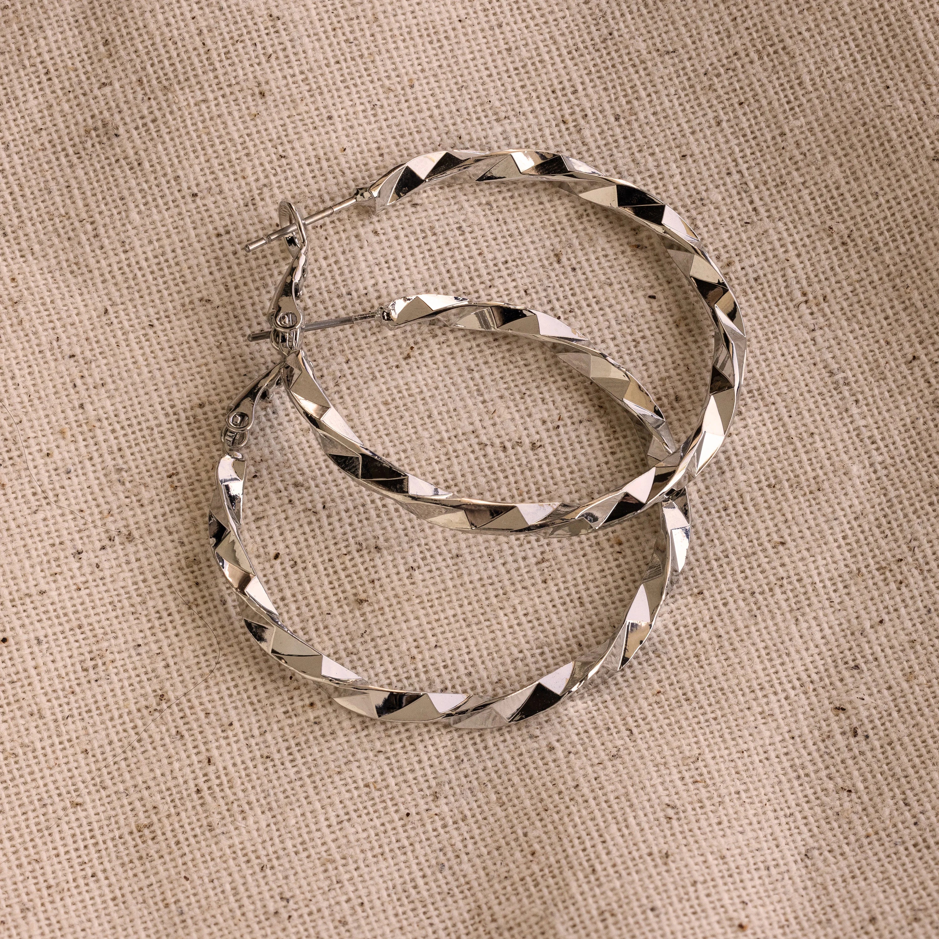The Large Twisted Hoops, in silver, rest on a beige woven fabric background—an ideal choice for summer style.