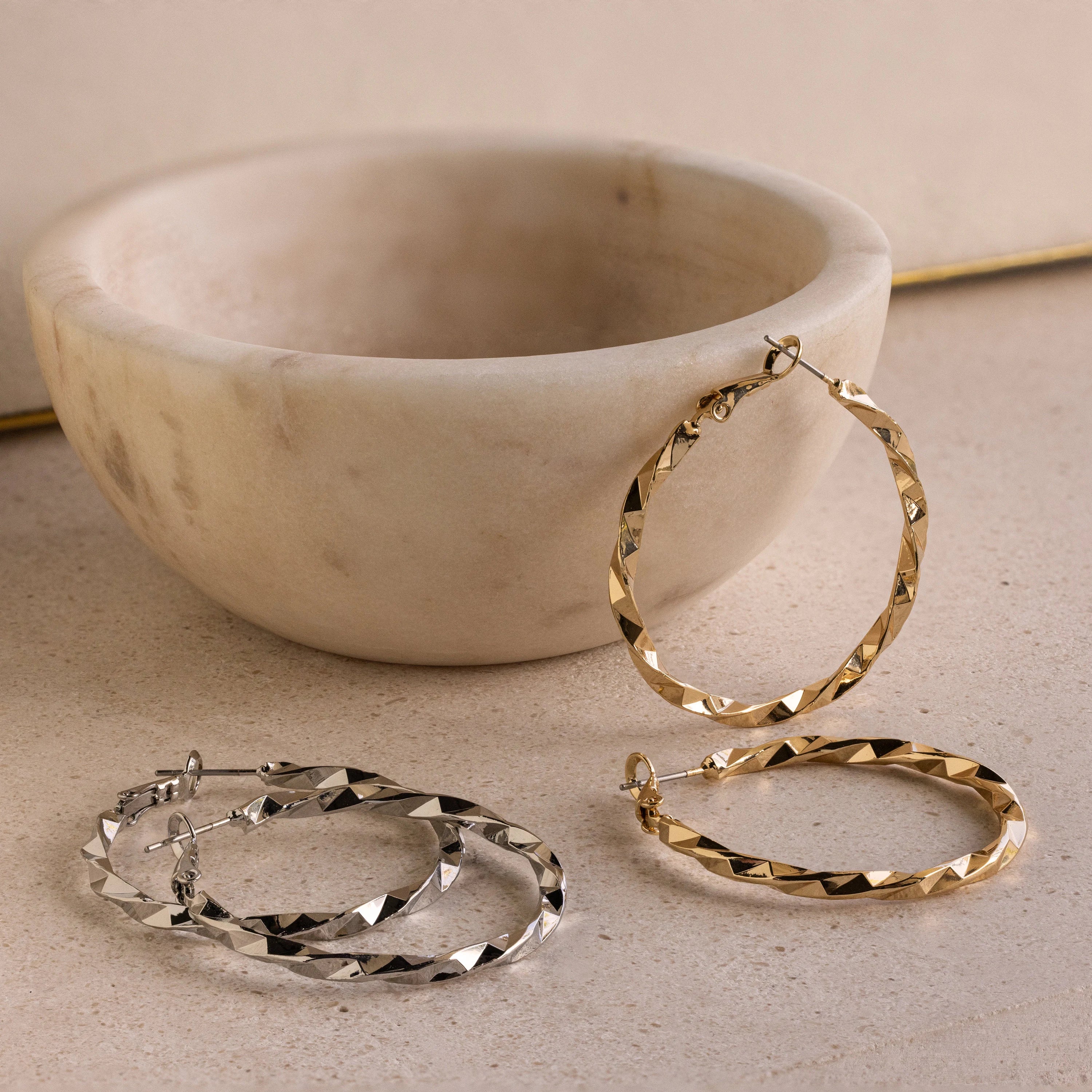 Large Twisted Hoops are displayed near a round, light-colored stone bowl—their bold, textured twist design makes them the perfect summer accessory.