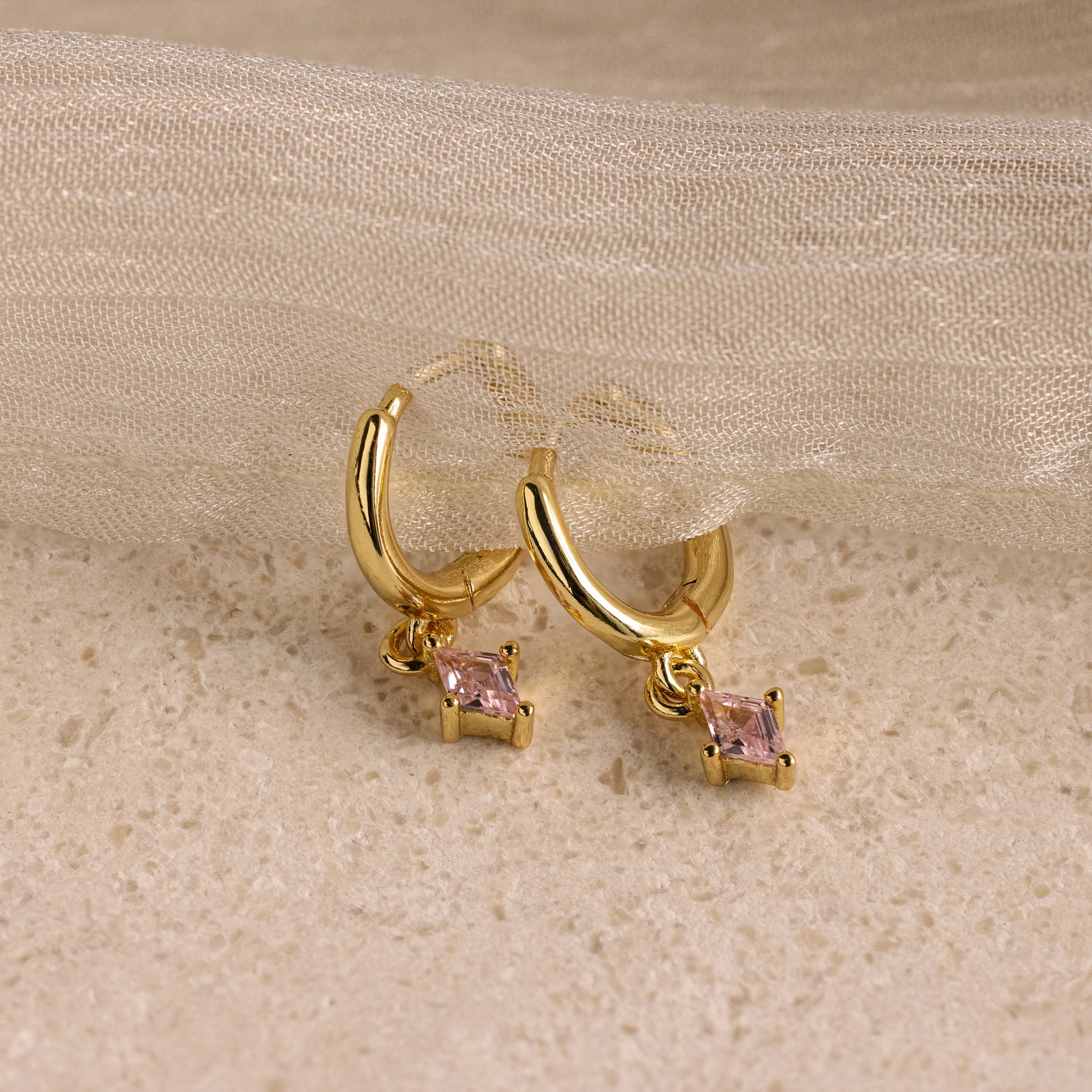 Birthstone Drop Huggies—gold huggie earrings with dangling pink gemstones, shown on beige textured fabric. These earrings are ideal personalized jewelry for adding a unique touch to your style.