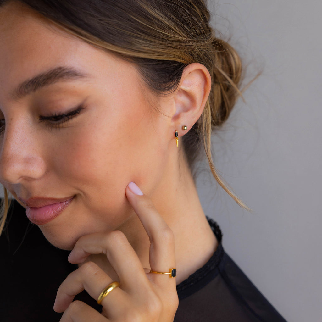 Woman wearing Onyx Dagger Drop Earrings and a black top, with her hair in a loose bun, touches her face.