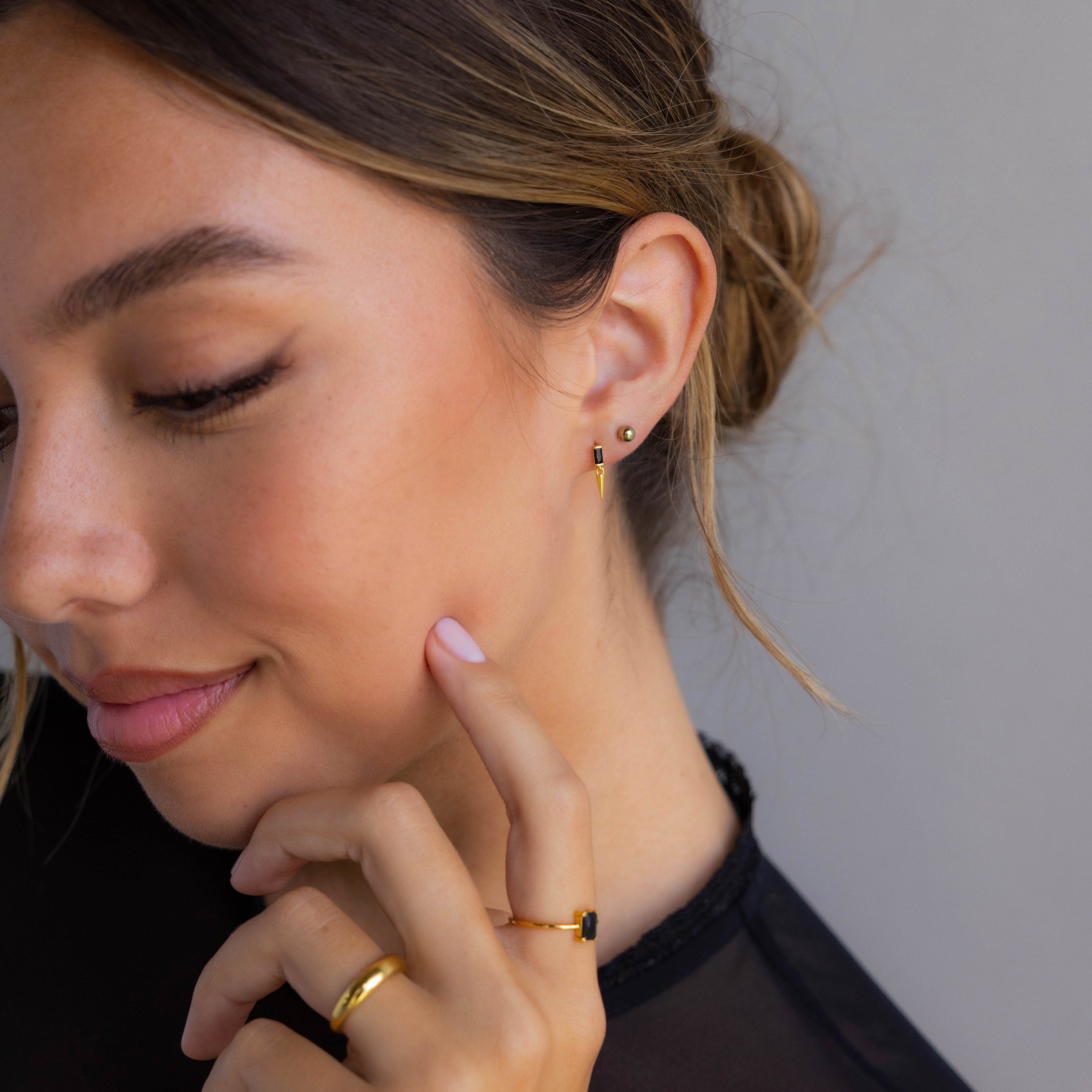 Woman wearing Onyx Dagger Drop Earrings and a black top, with her hair in a loose bun, touches her face.
