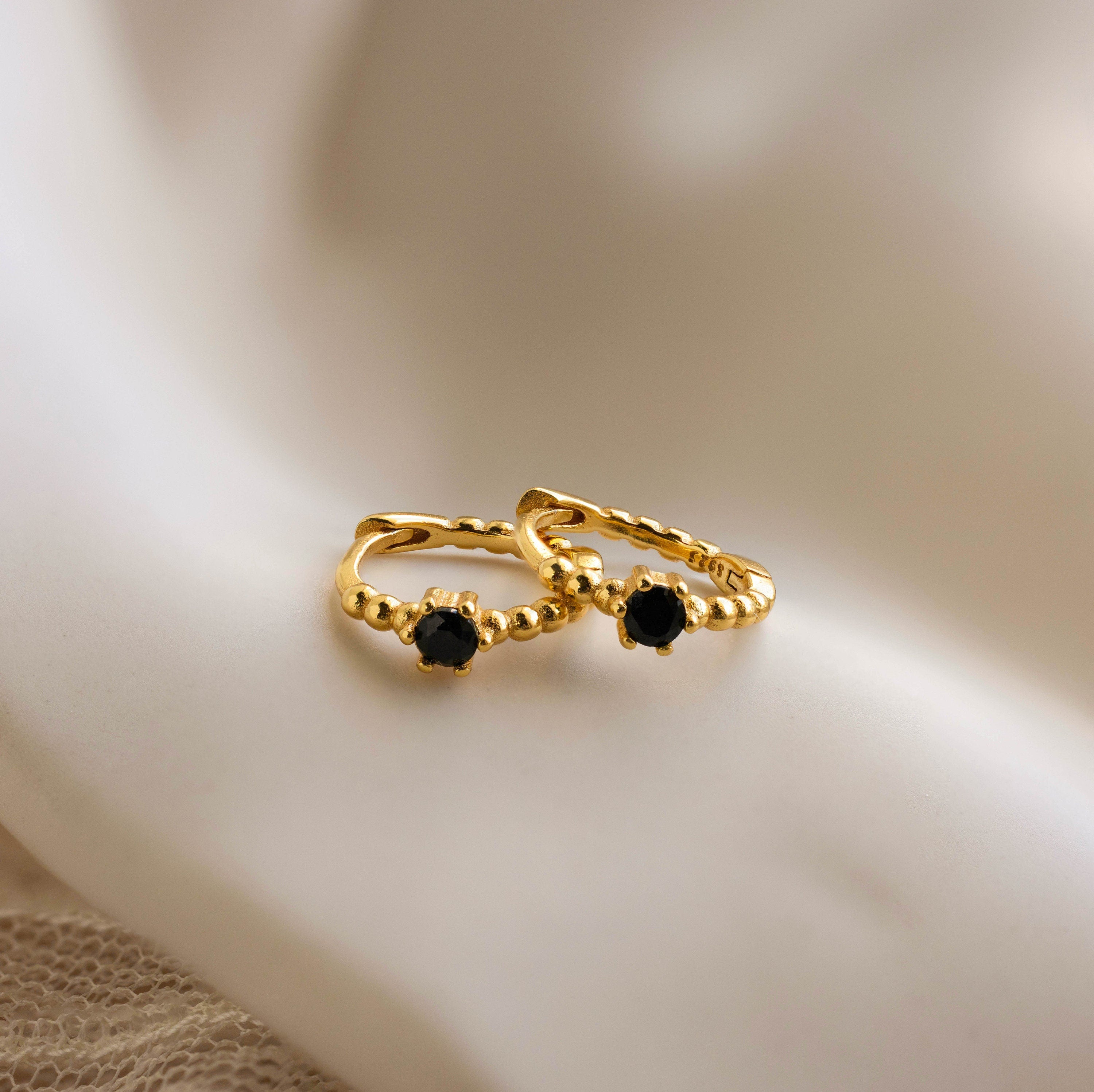 Two small Onyx Beaded Huggies with black gemstones, displayed on a soft, white fabric background.