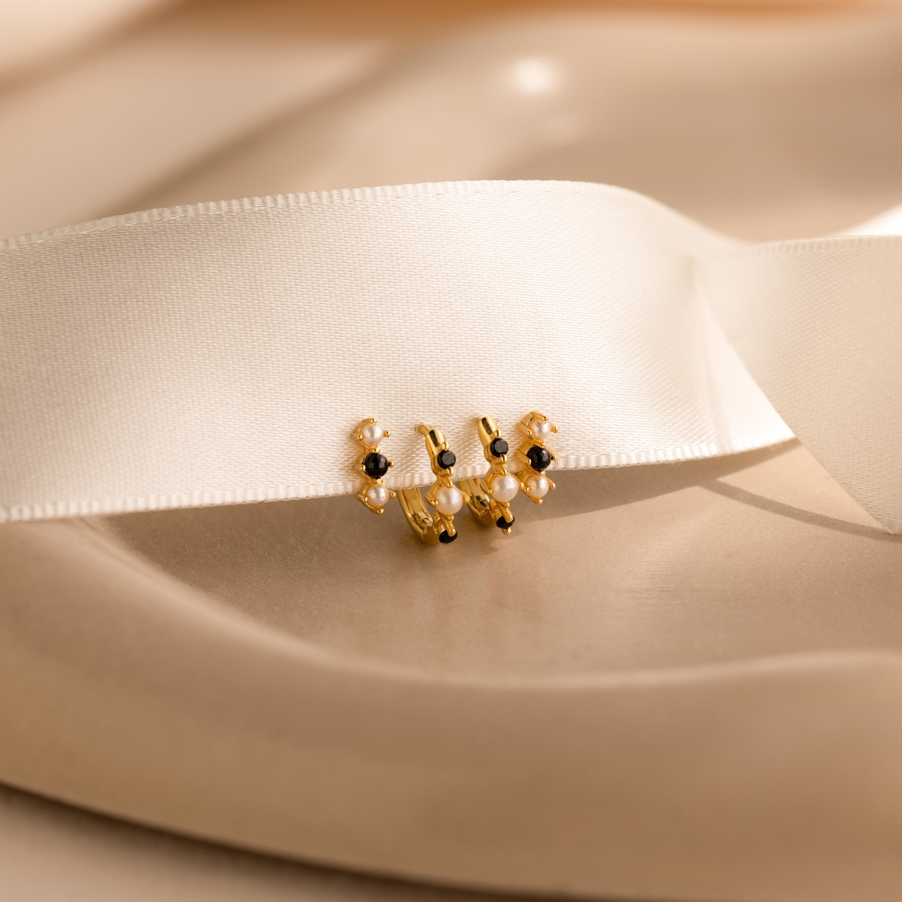 Pearl & Onyx Huggies, gold and black beaded earrings inspired by elegant pearls, are displayed on a white satin ribbon with a soft beige background.