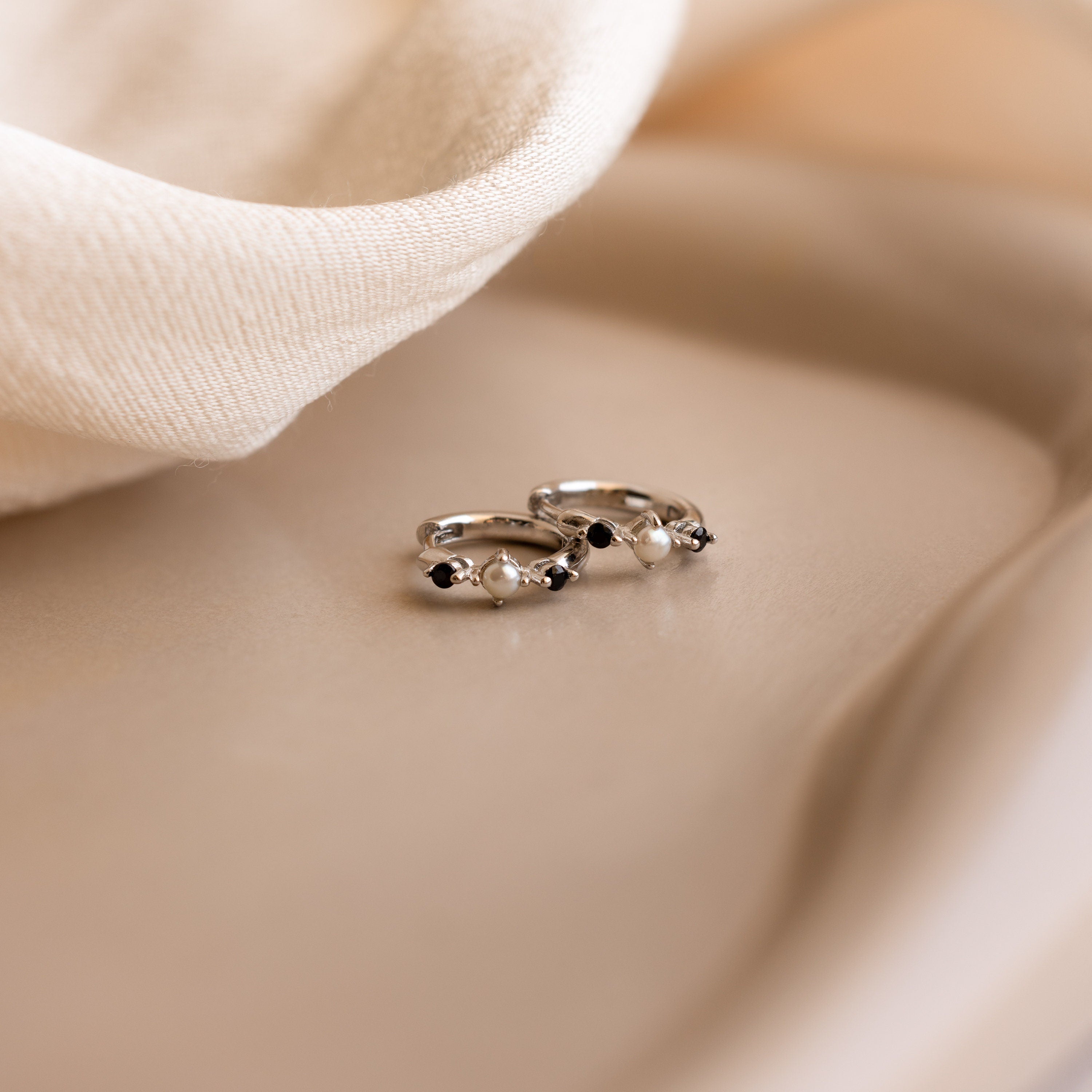 Two silver Pearl & Onyx Huggies, adorned with small black stones and pearls, rest on a soft beige fabric background.