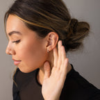 A woman with brown hair in a bun holds her hand to her ear as if listening, her Pearl & Onyx Huggies bringing an elegant touch to the moment.