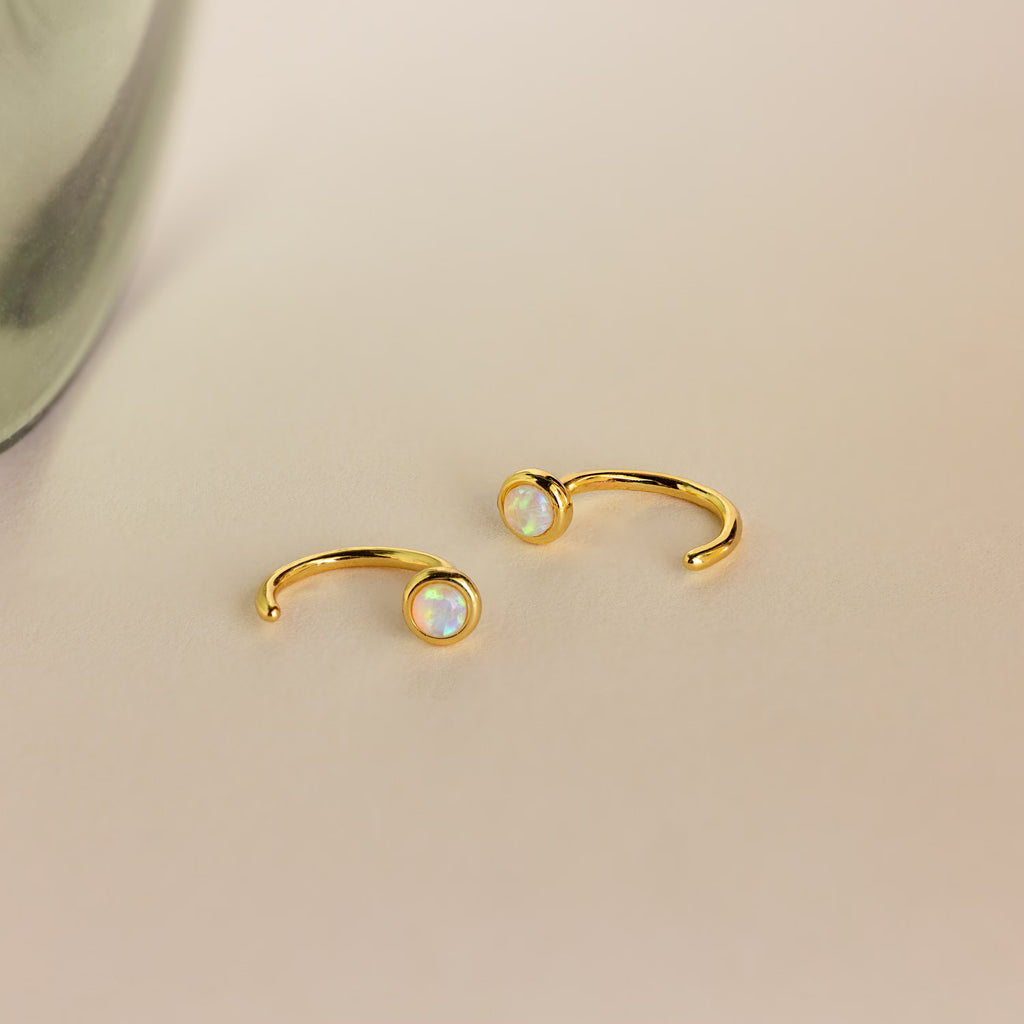 The Opal Threader Huggies, gold open rings with round opal stones and celestial inspiration, rest gracefully on a light beige surface.
