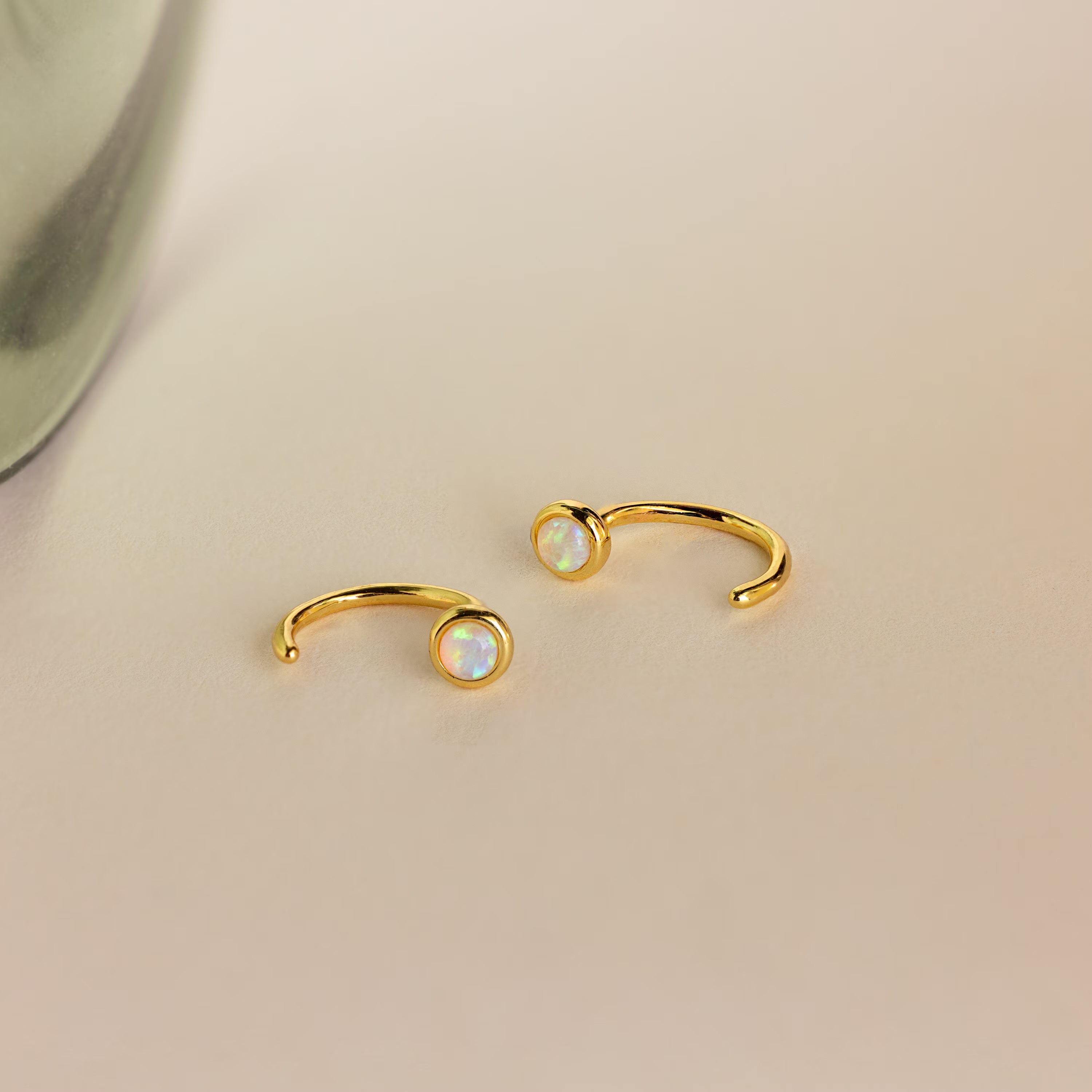 The Opal Threader Huggies, gold open rings with round opal stones and celestial inspiration, rest gracefully on a light beige surface.