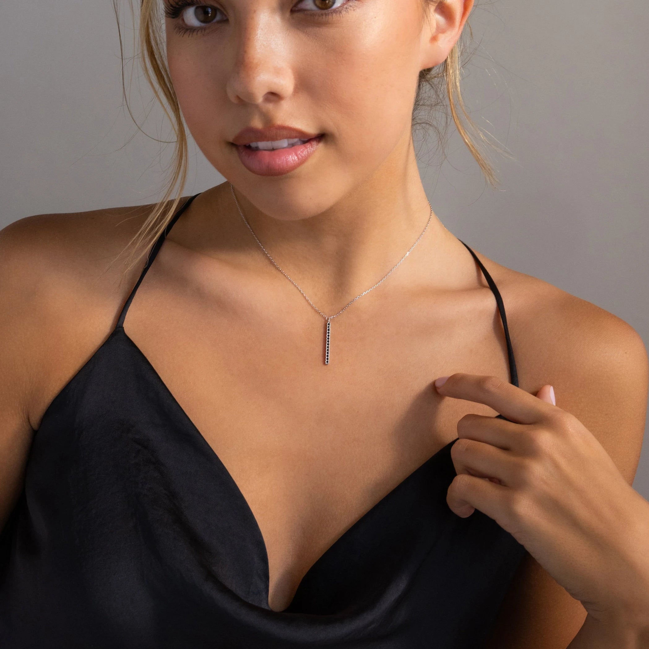 A woman in a black satin dress touches her collarbone while wearing the Onyx Bar Pendant Necklace, featuring a sleek modern bar design.