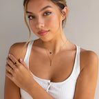 A woman in a white tank top showcases gold jewelry, featuring the Onyx Bar Pendant Necklace and matching onyx gemstone rings, while posing against a neutral background.