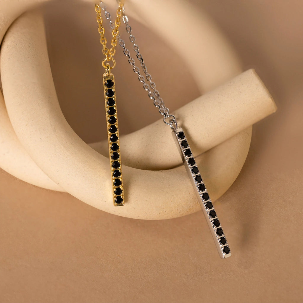 Two Onyx Bar Pendant Necklaces, one in gold and one in silver, each featuring a striking onyx gemstone, are elegantly displayed on a beige sculpture.
