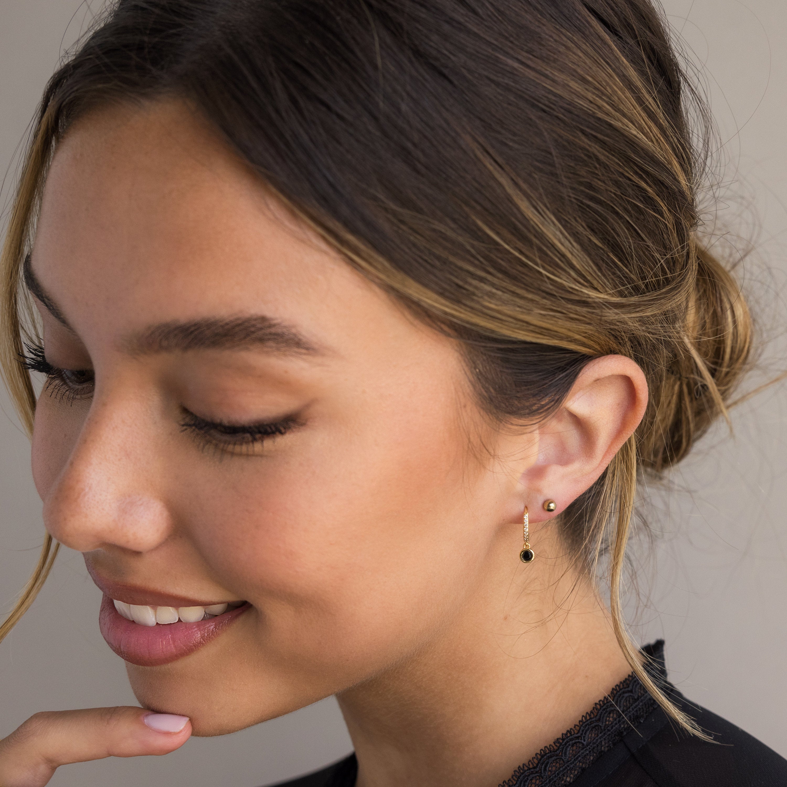 A woman with a low bun smiles, gently touching her chin. She wears a black top and the elegant Pave Onyx Drop Huggies—small gold earrings adorned with subtle black onyx gemstones for a refined, sophisticated touch.