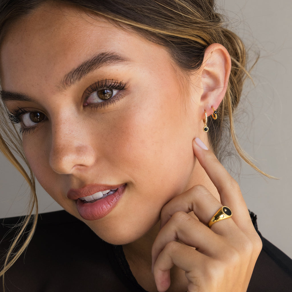 A woman with natural makeup wears Pave Onyx Drop Huggies and a gold ring, touching her ear as she looks at the camera.