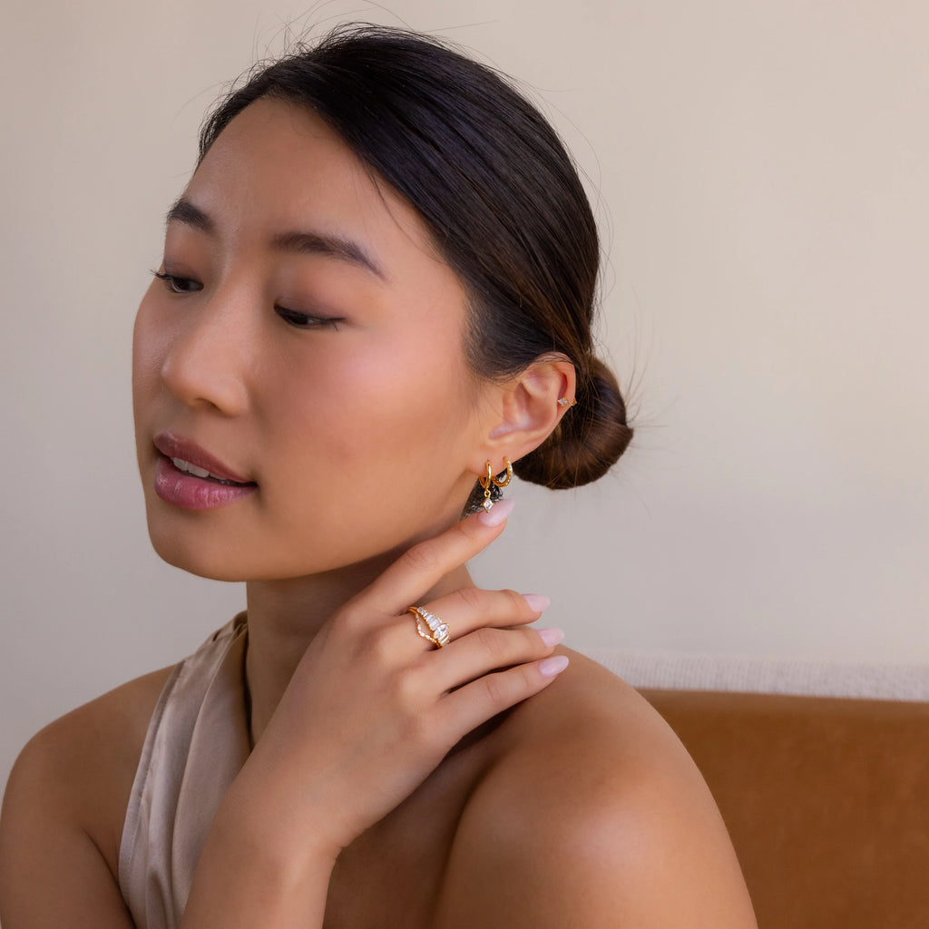 A woman with dark hair in a low bun wears the Diamond Pave Earrings Set and gold jewelry, gently touching her neck as she gazes down thoughtfully.