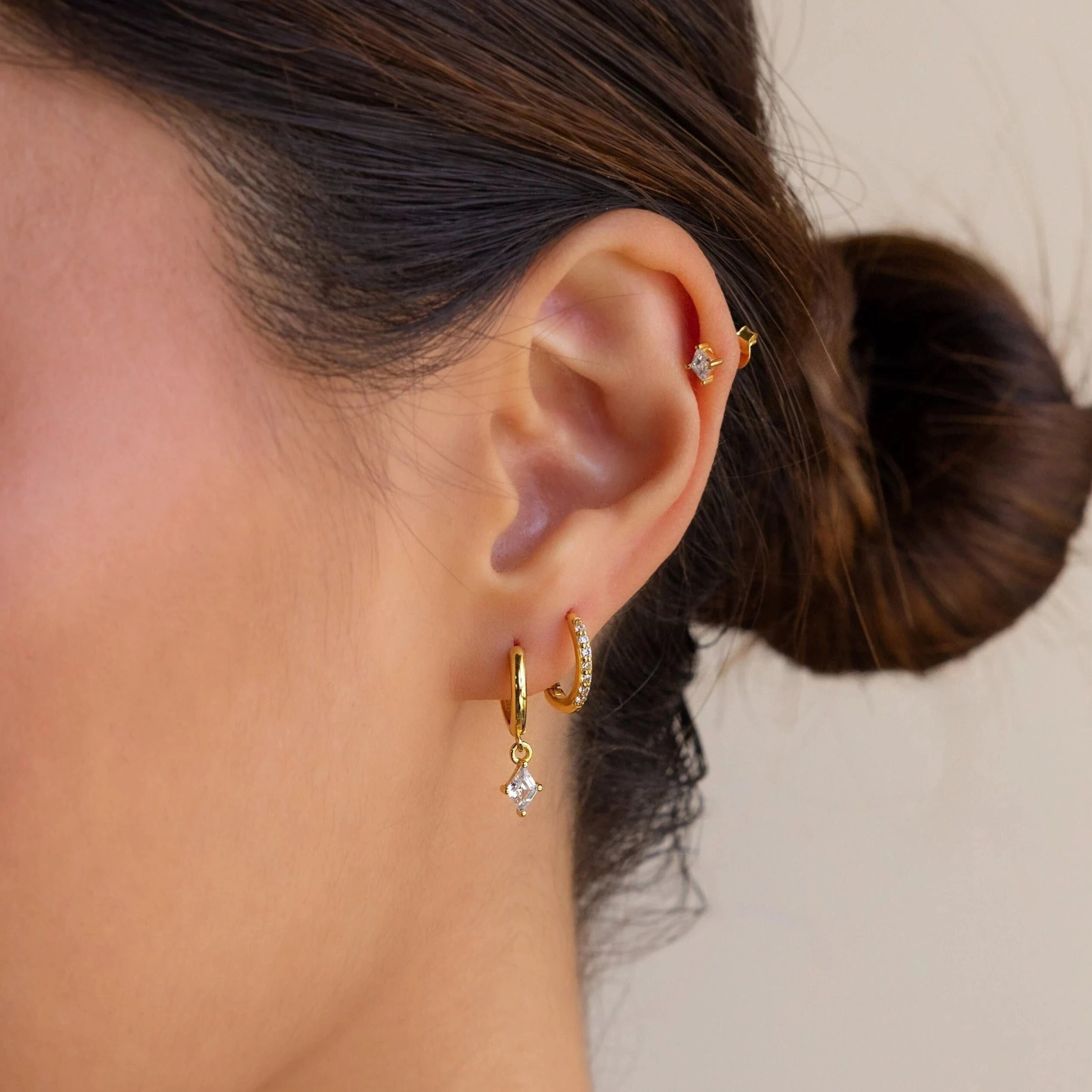 A womans ear with hair in a bun showcases the Diamond Pave Earrings Set—featuring gold and sterling silver small hoops and a diamond pave earring—against a neutral background for a chic bridal look.