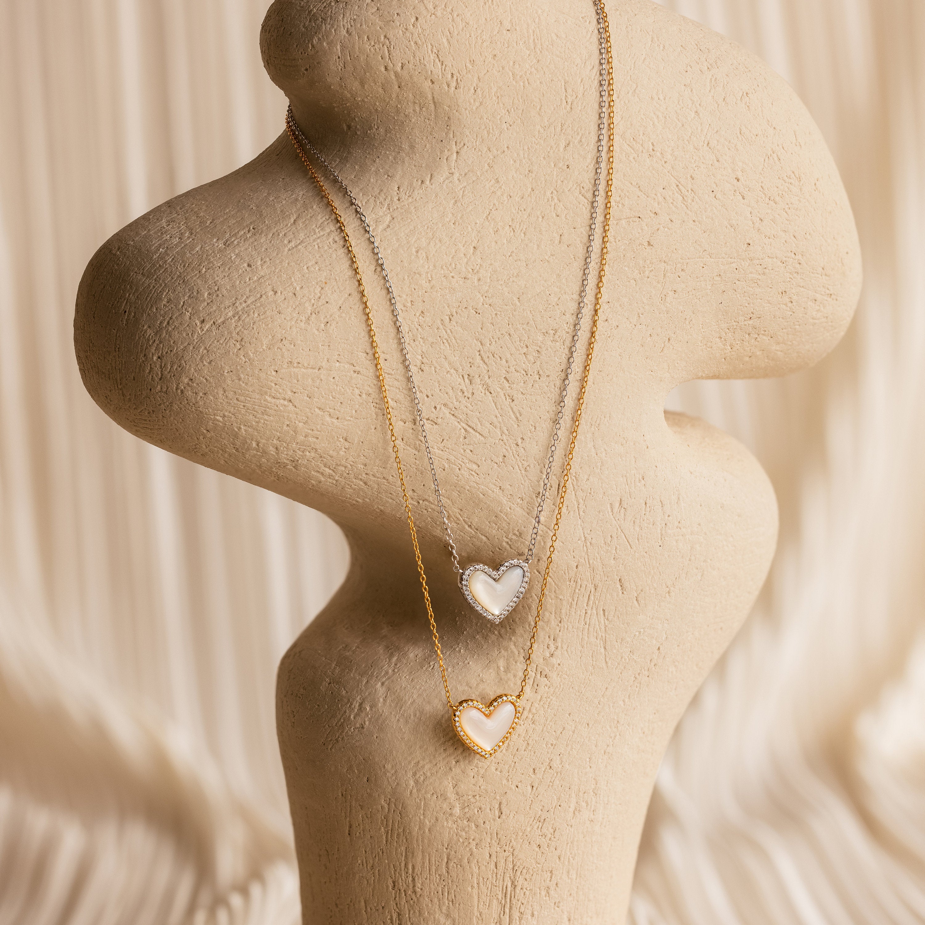 The Pave Mother of Pearl Heart Necklace, available in silver and gold, is showcased on a beige abstract stand—a timeless duo ideal for those who love heirloom-inspired jewelry.