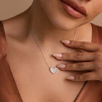 A woman wearing the Pave Mother of Pearl Heart Necklace with shimmering silver, paired with a brown satin top, gently touches her collarbone.