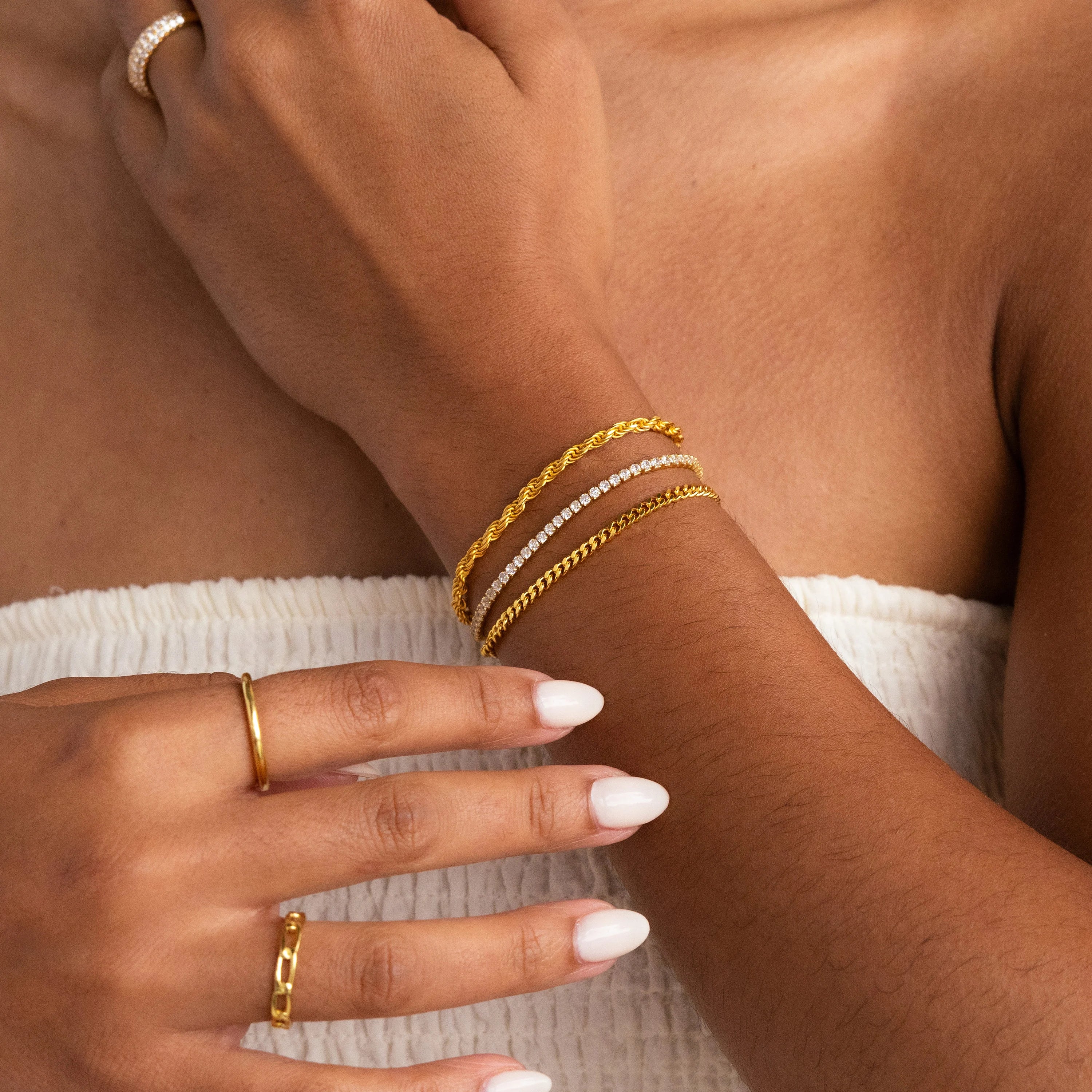 A woman with manicured nails and a white textured strapless top showcases a chic layered look, styled with gold rings and the Curb Chain Bracelet.
