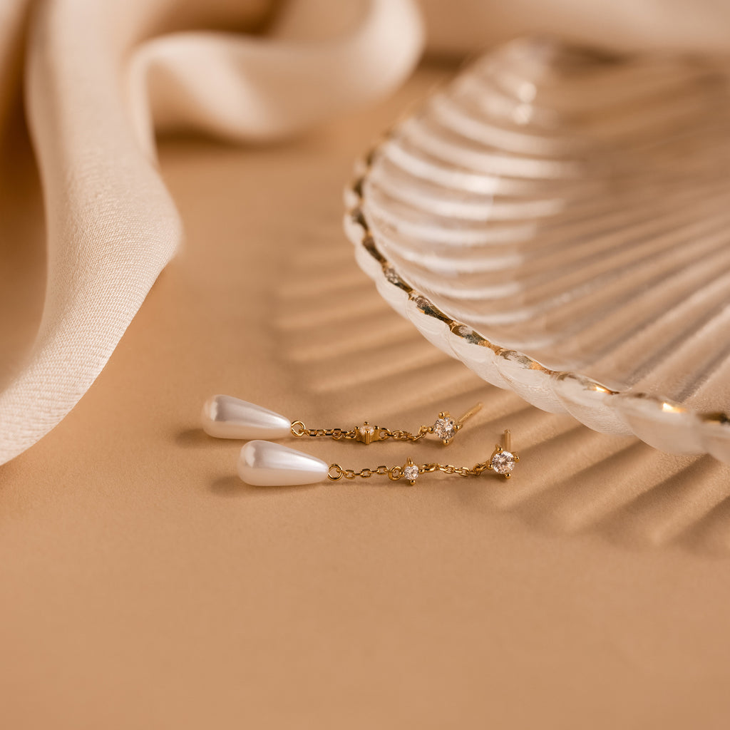 Diamond Pearl Chain Earrings with a pearl teardrop charm sit beside a glass shell dish on beige fabric.