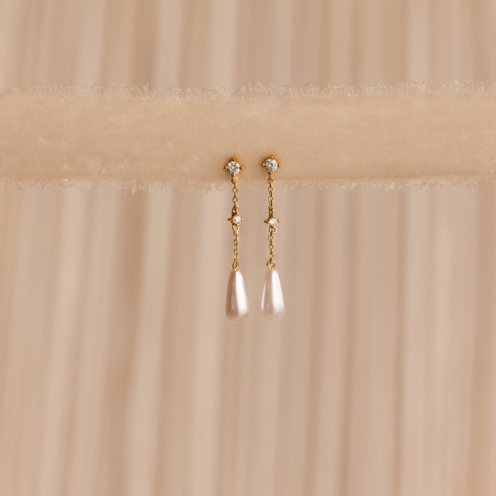 Diamond Pearl Chain Earrings are elegantly displayed on a soft beige fabric background, showcasing their sparkling studs and delicate teardrop pearl charm.