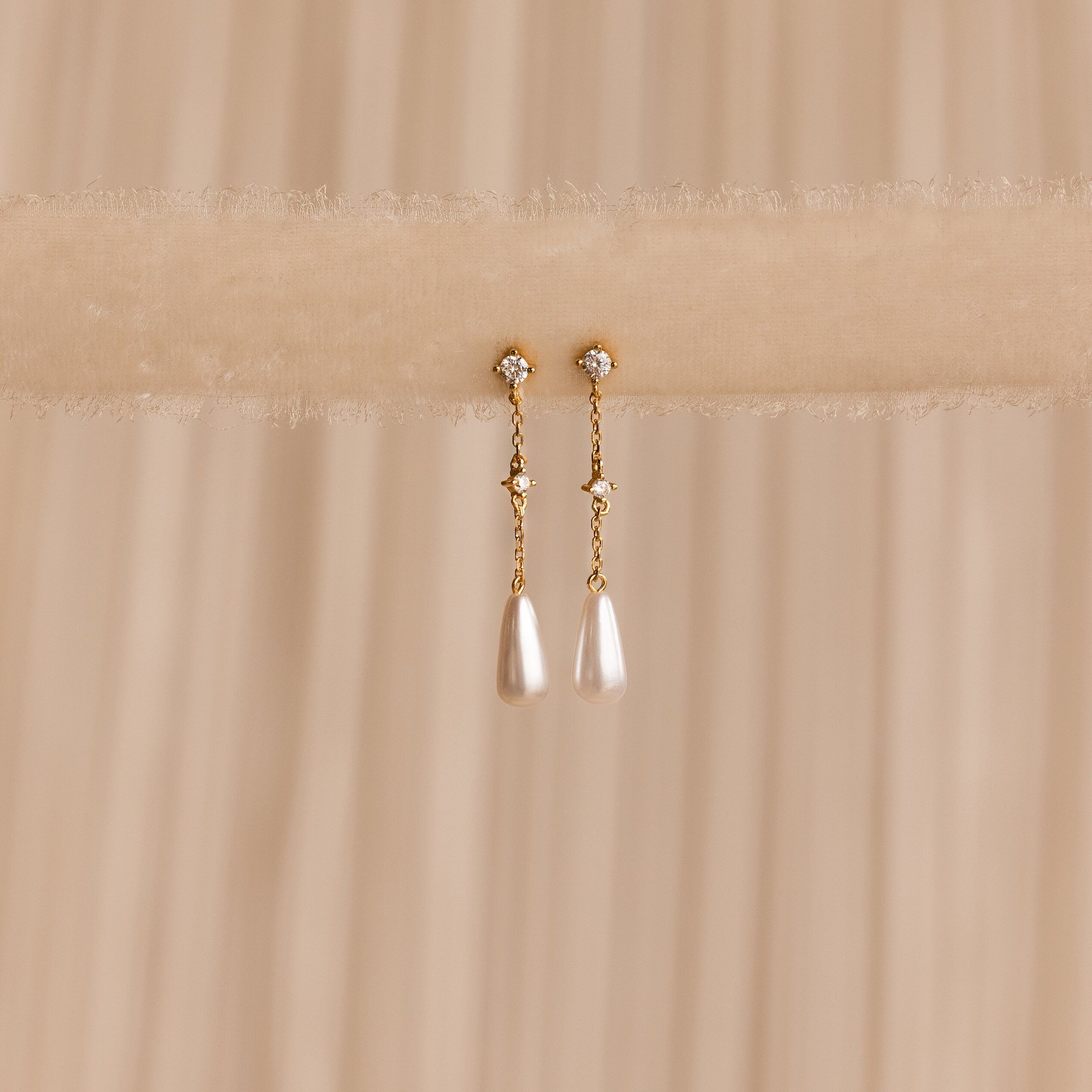Diamond Pearl Chain Earrings are elegantly displayed on a soft beige fabric background, showcasing their sparkling studs and delicate teardrop pearl charm.