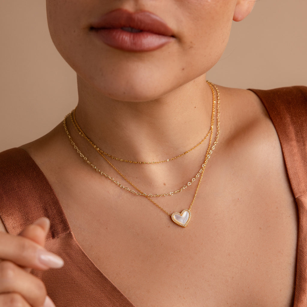Woman wearing layered gold necklaces, featuring the Pave Mother of Pearl Heart Necklace, styled with a brown satin top.