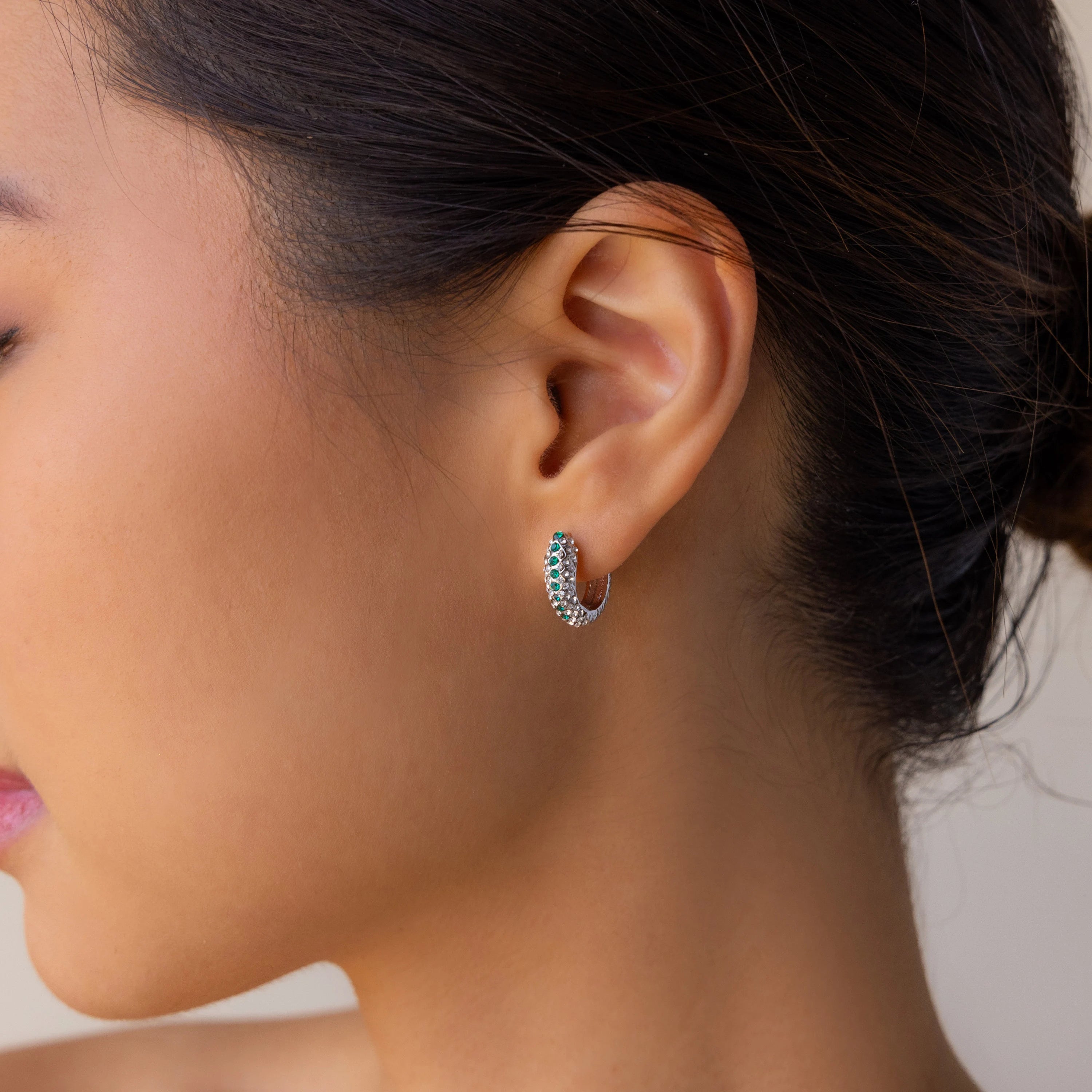 A woman shown from the side wears Emerald Pave Snake Hoops, silver earrings inspired by snakeskin texture, set with sparkling stones, against a neutral background.