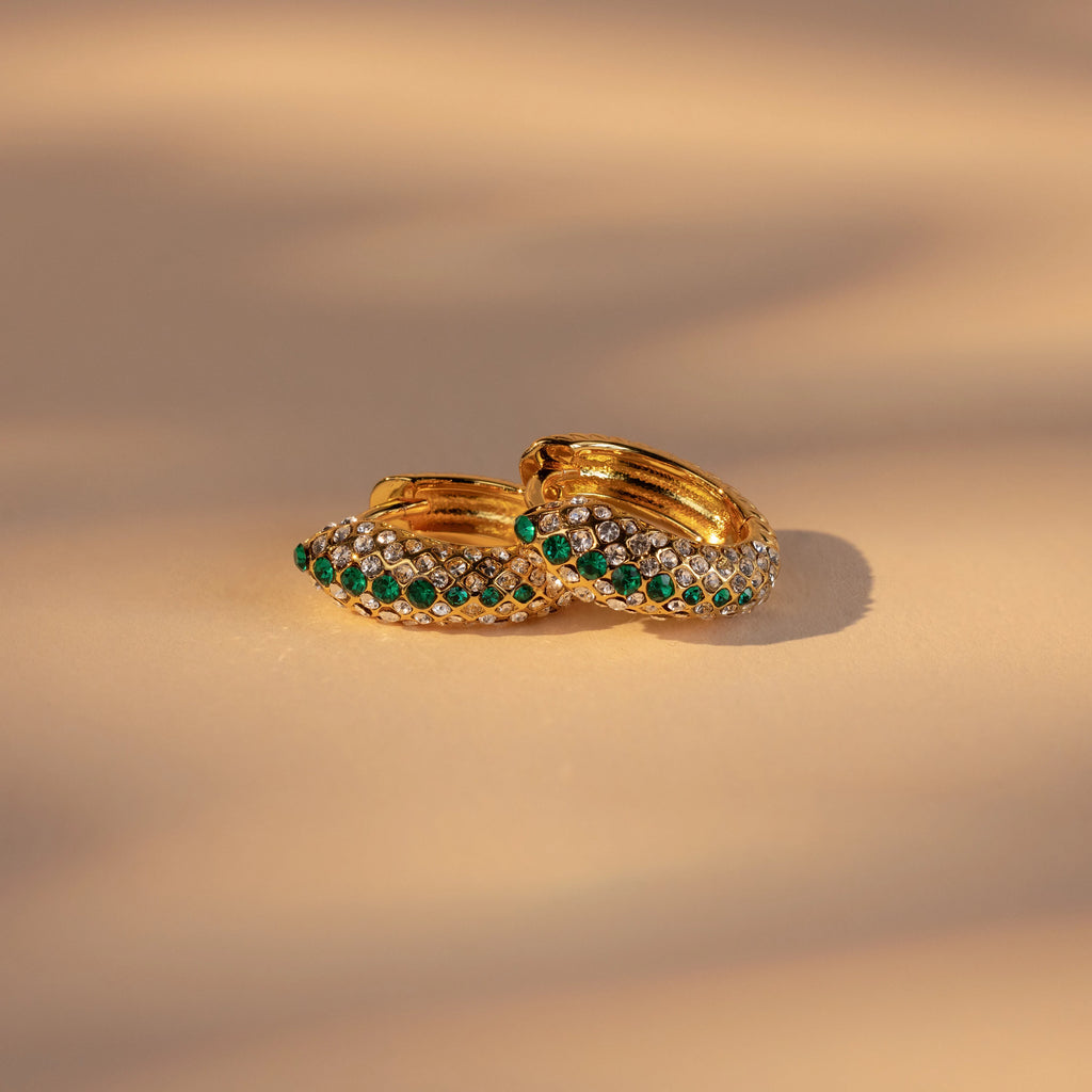 Emerald Pave Snake Hoops—gold hoops adorned with green and white gemstones—are shown on a beige surface under soft lighting.