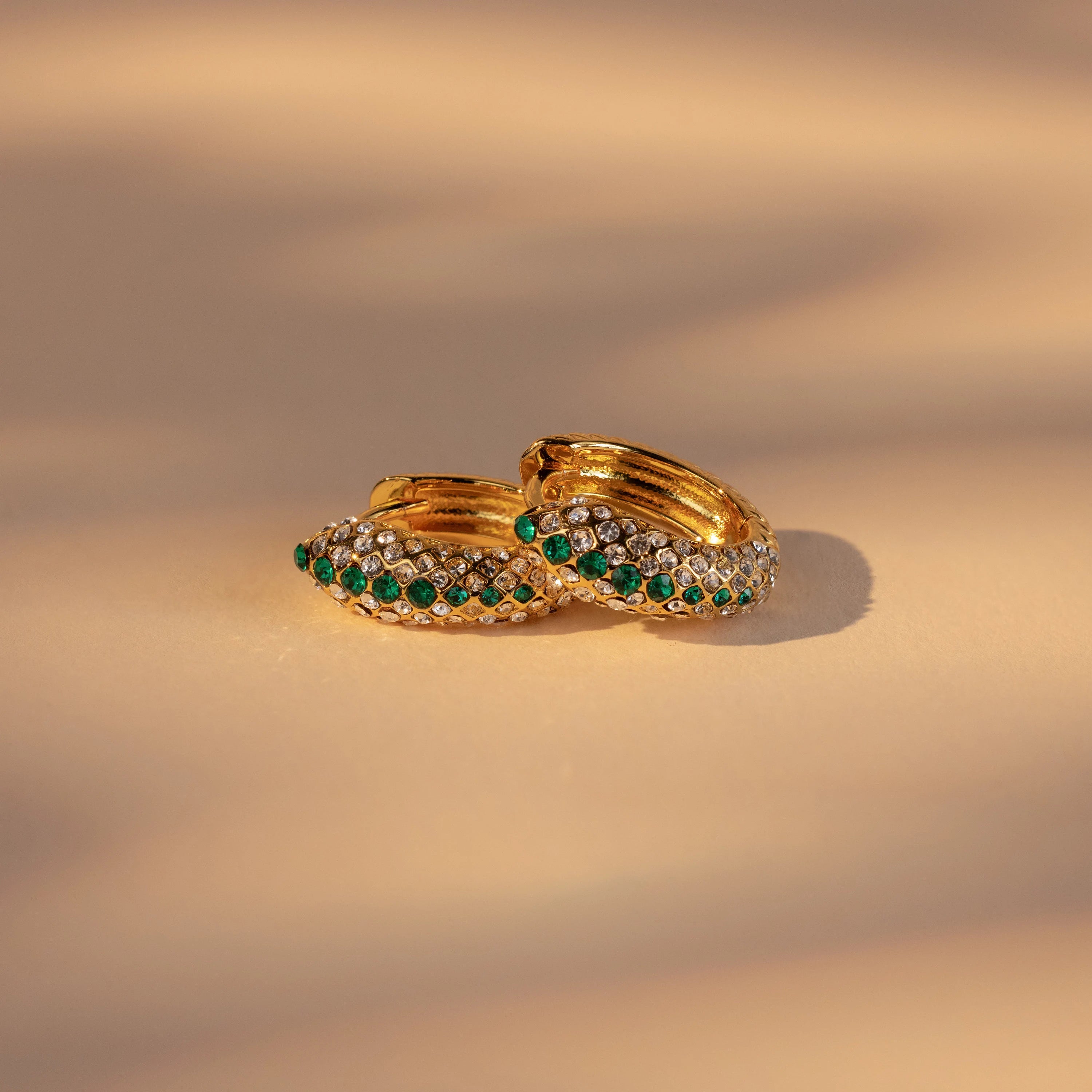 Emerald Pave Snake Hoops—gold hoops adorned with green and white gemstones—are shown on a beige surface under soft lighting.