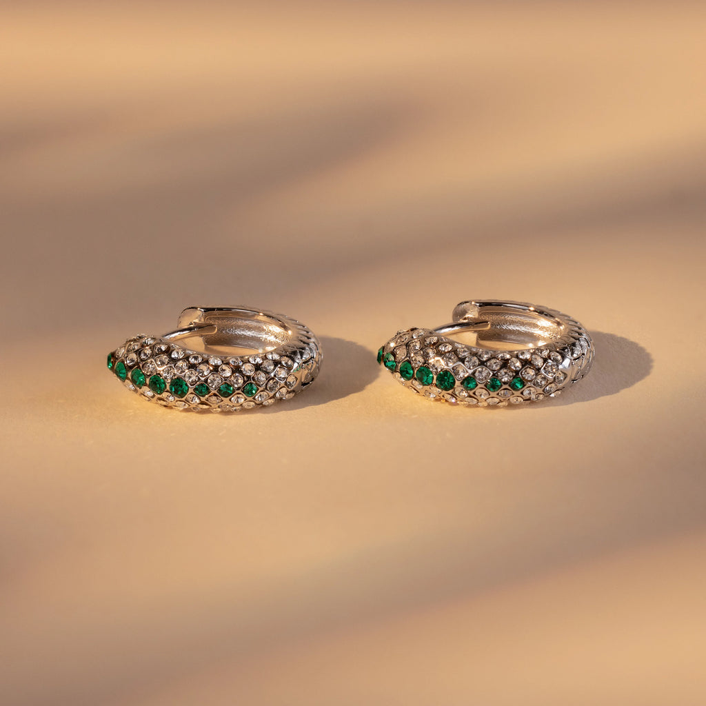 The Emerald Pave Snake Hoops are gold hoop earrings with green and clear gemstones and a subtle snakeskin texture, shown on a beige background.