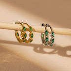 The Emerald Marquise Inlay Huggies, featuring gold and silver with green gemstones, are elegantly displayed on a wooden rod against a beige background—ideal for fans of dainty jewelry.