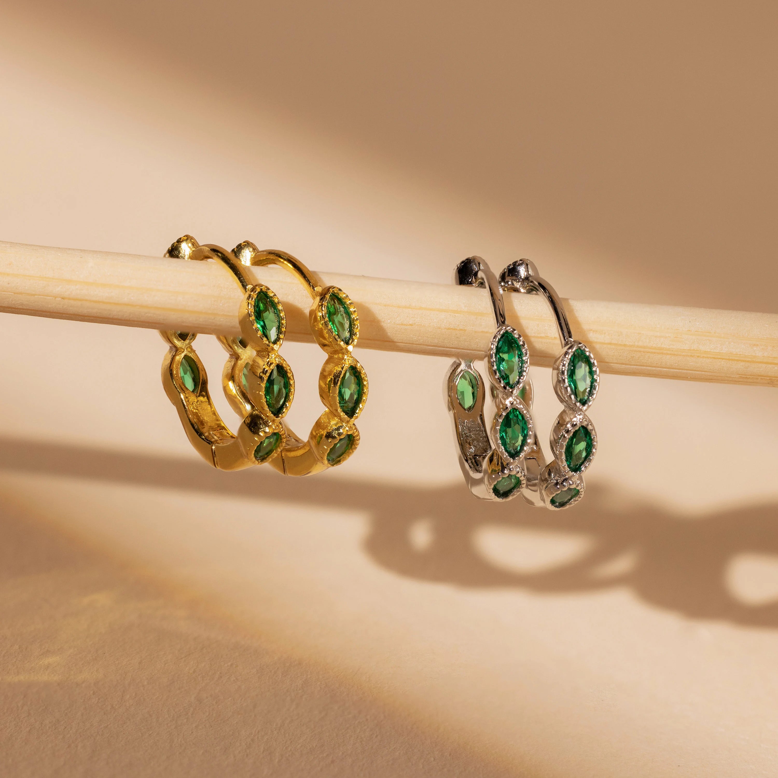 The Emerald Marquise Inlay Huggies, featuring gold and silver with green gemstones, are elegantly displayed on a wooden rod against a beige background—ideal for fans of dainty jewelry.