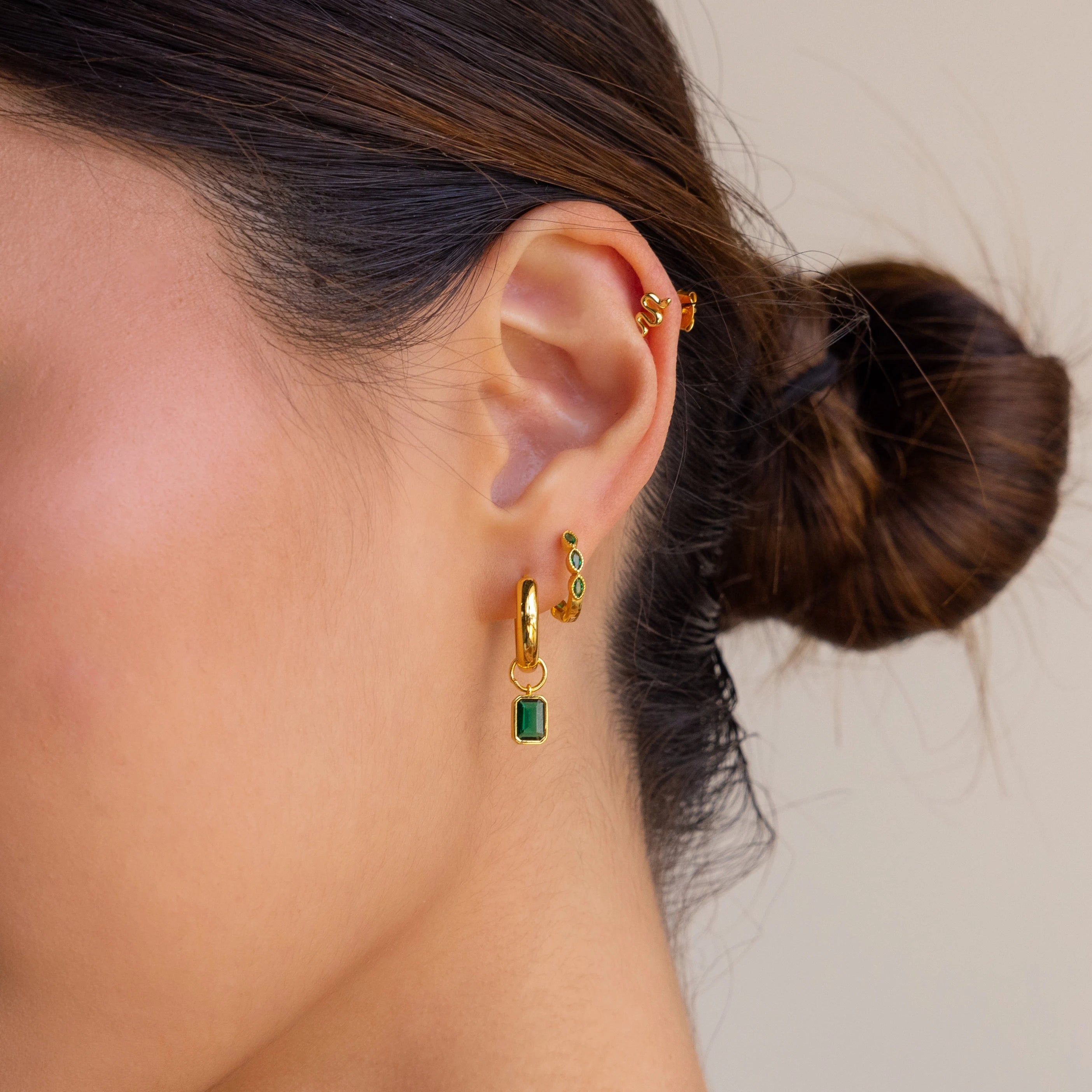 A woman's ear adorned with Emerald Marquise Inlay Huggies and small gold studs, her hair styled in a low bun.