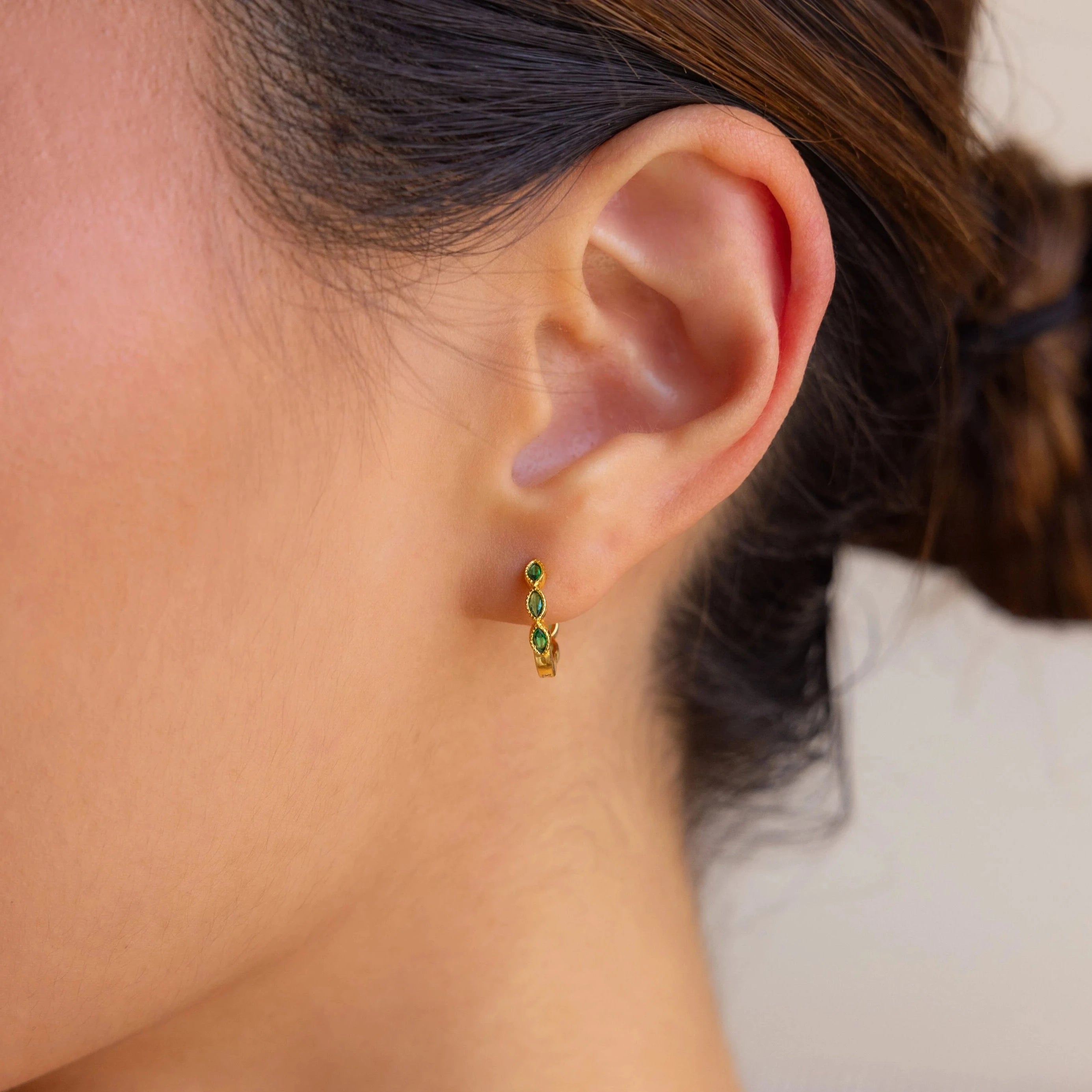 A close-up of a woman's ear showcases the Emerald Marquise Inlay Huggies, featuring dainty emerald accents that add a touch of sophistication.