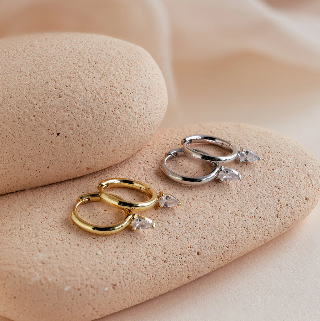 The Pear Diamond Drop Hoops—available in gold and silver, each set with a teardrop-shaped stone—rest on textured stones, perfect as a wedding accessory or a chic addition to your elegant earring collection.
