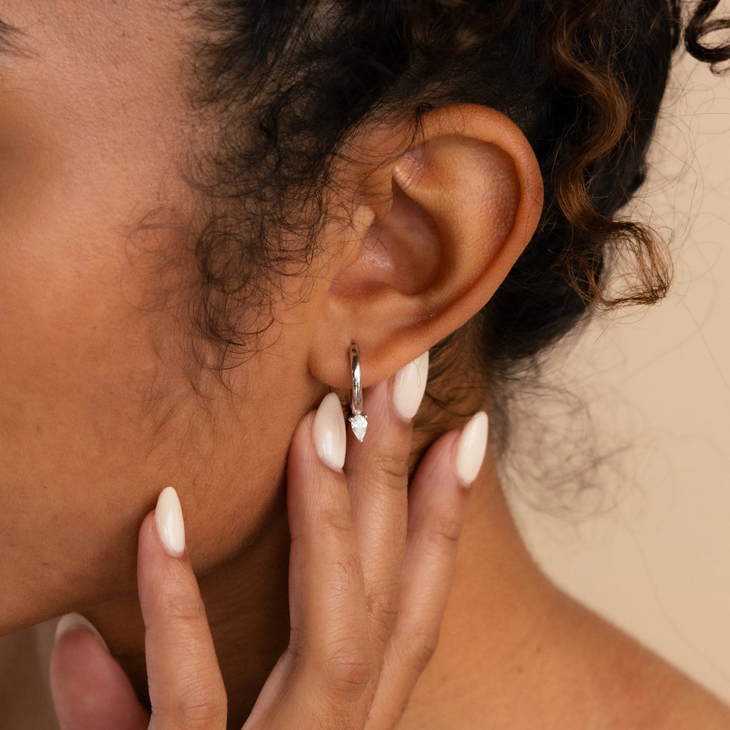 A close-up of a woman touching her ear, wearing elegant Pear Diamond Drop Hoops—silver earrings with a small dangling gem, perfect as a wedding accessory.