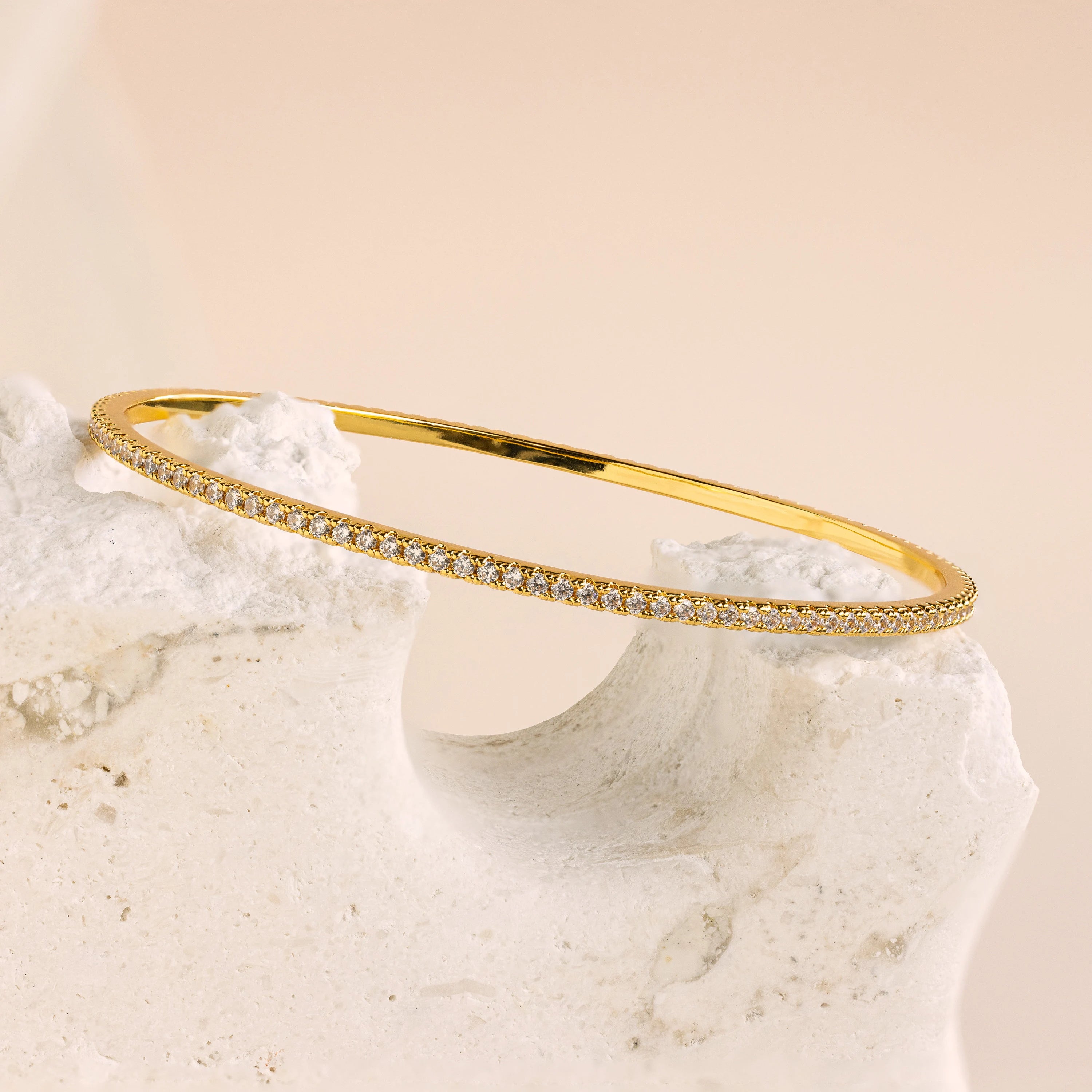 The Classic Diamond Tennis Bangle, crafted in gold with pavé stones and small diamonds, rests elegantly on a textured white stone.