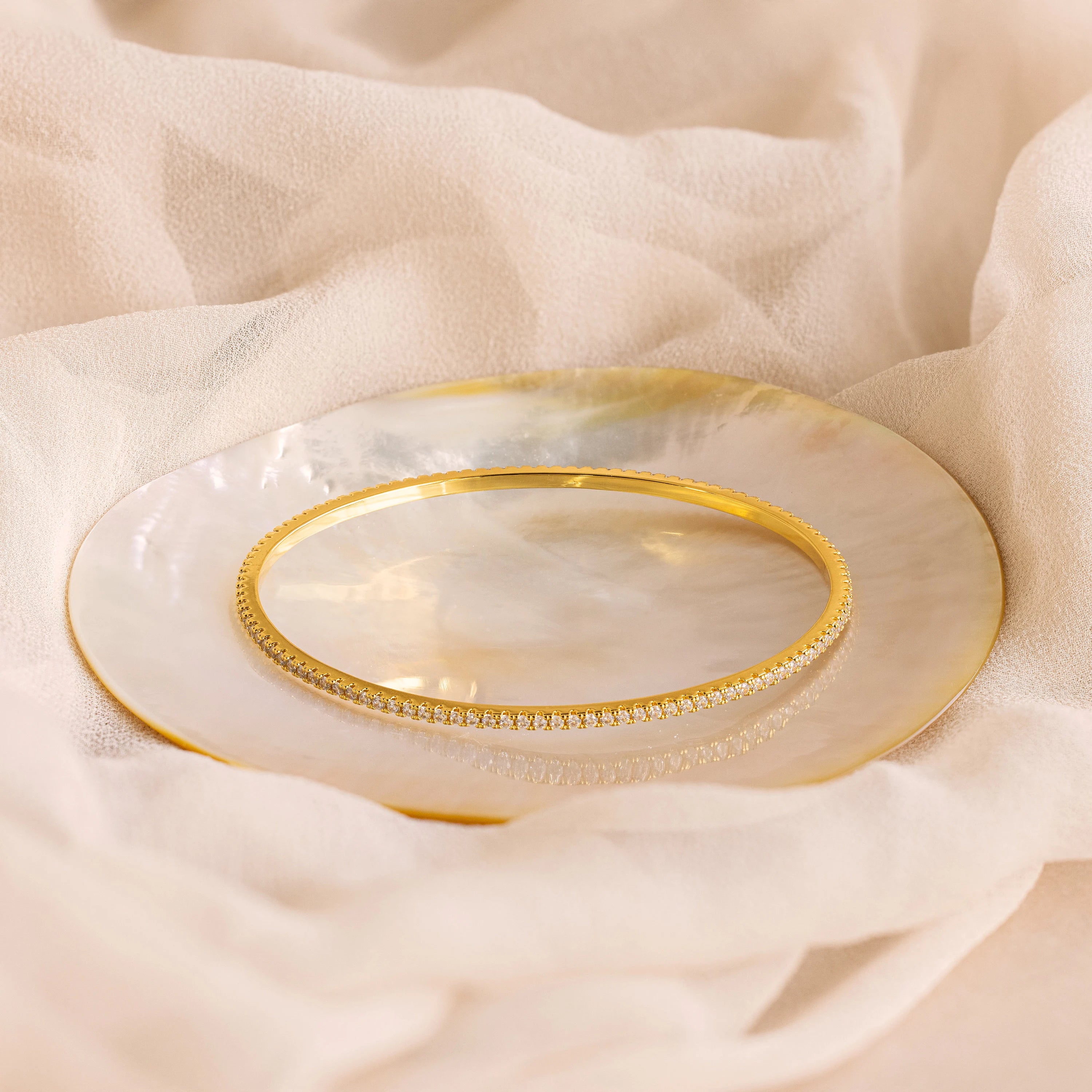 The Classic Diamond Tennis Bangle, accented with pavé stones, is displayed on a gold-rimmed mother-of-pearl plate set against soft beige fabric.