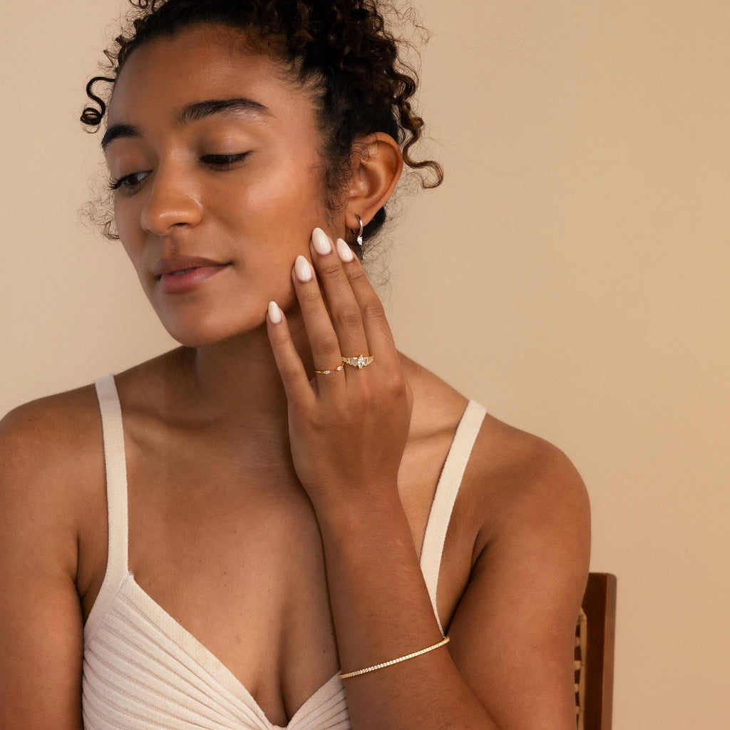 A woman in a beige top touches her face, showing off elegant earrings and white manicured nails. The Pear Diamond Drop Hoops shimmer beautifully as a wedding accessory against the neutral background.
