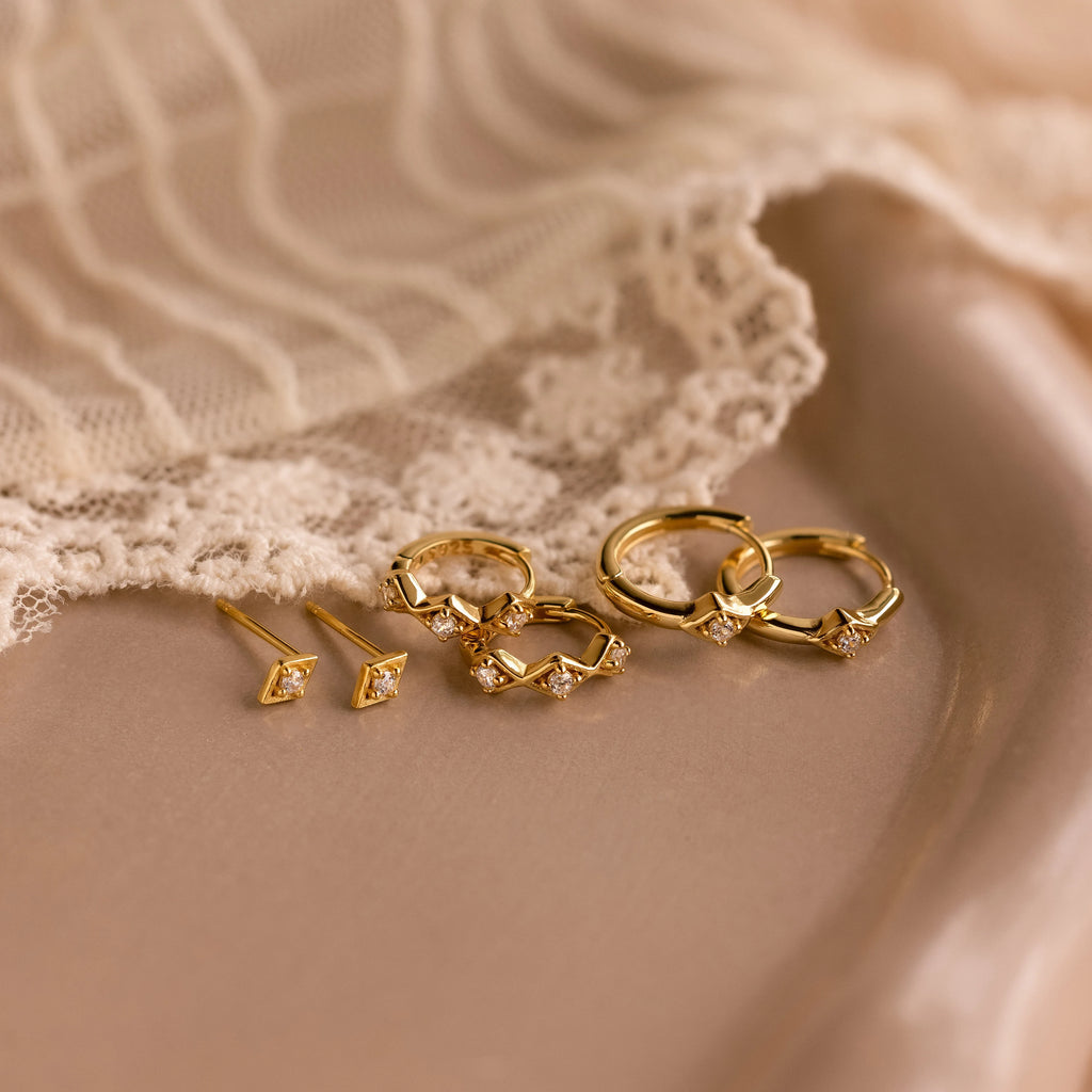 The Ericka Diamond Tennis Huggies, elegant gold earrings with small diamonds, are displayed on a beige surface next to delicate lace fabric.