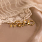 The Ericka Diamond Tennis Huggies, elegant gold earrings with small diamonds, are displayed on a beige surface next to delicate lace fabric.