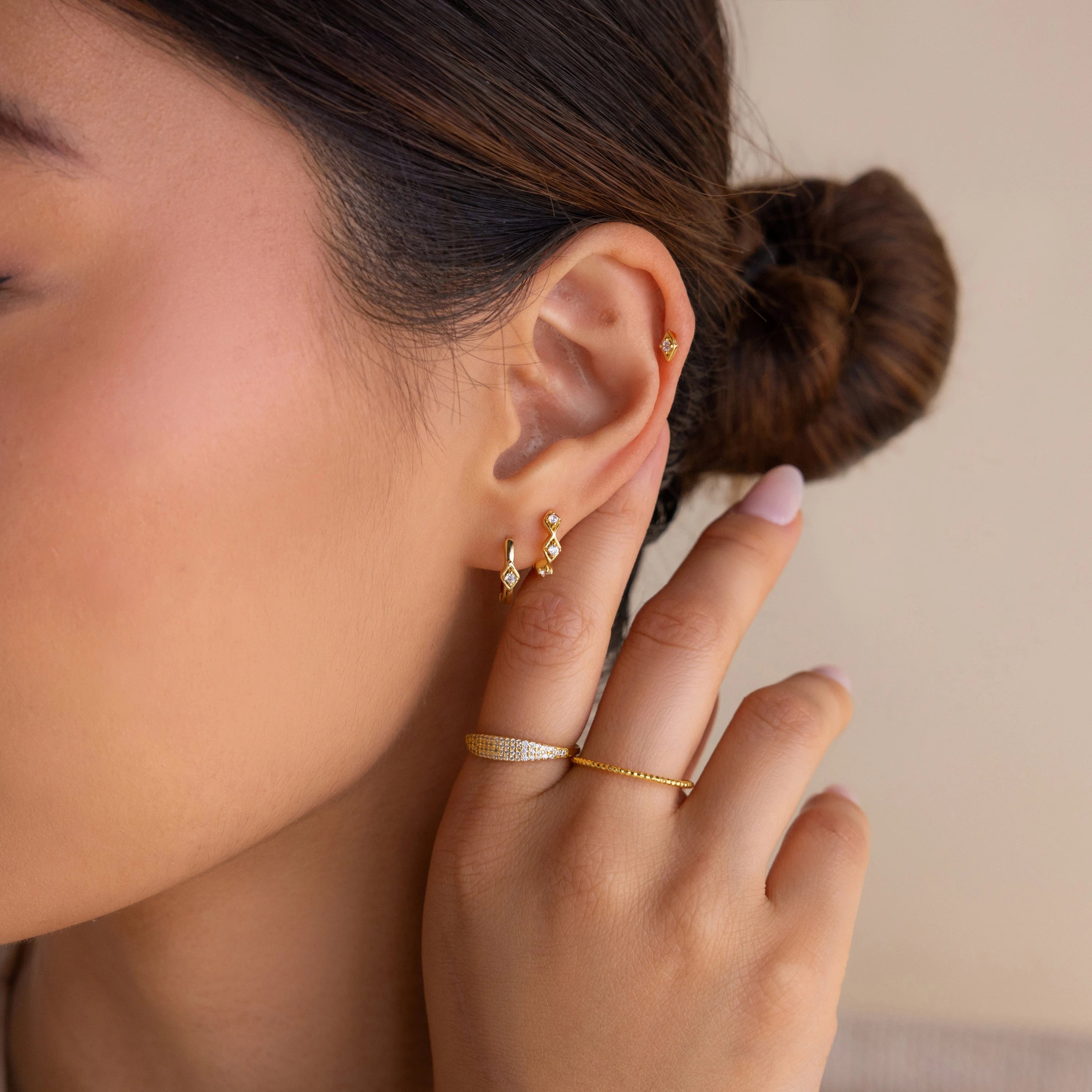 A woman with a low bun wears Ericka Diamond Tennis Huggies, a gold ring, and touches her ear with manicured fingers.