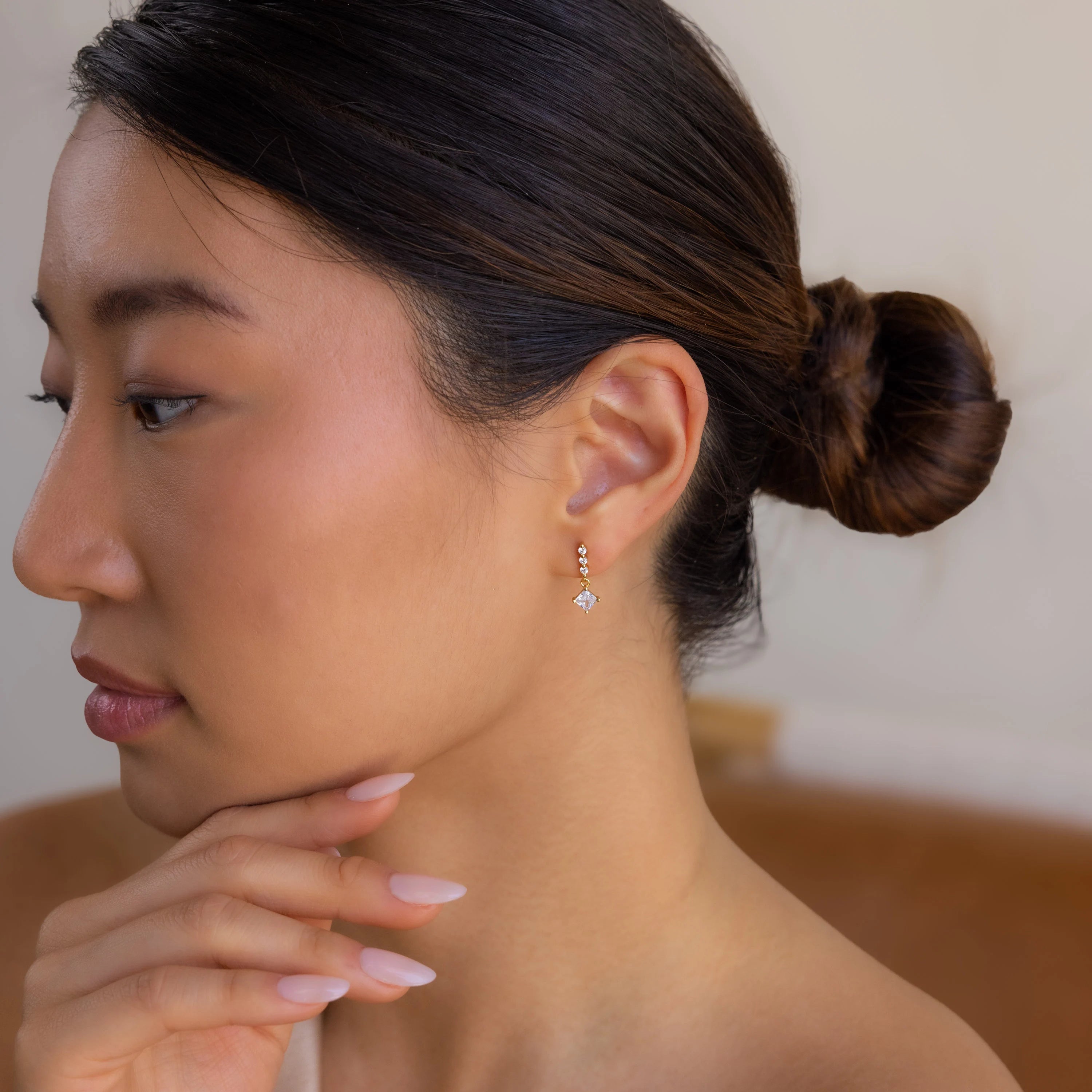 A woman with a neat bun wears Princess Diamond Drop Studs, delicately resting her chin on her hand.