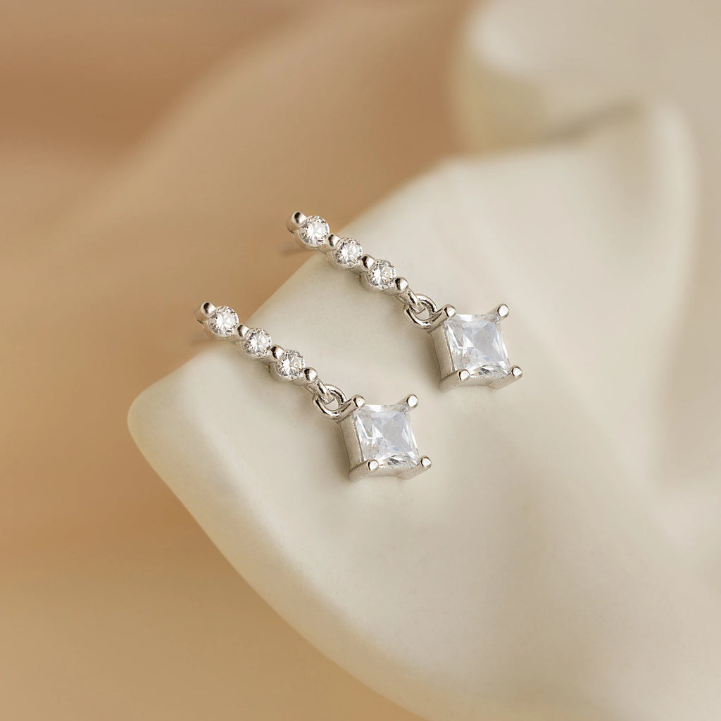 Princess Diamond Drop Studs featuring square princess cut gemstones and round clear stones displayed on a cream-colored background.