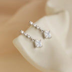 Princess Diamond Drop Studs featuring square princess cut gemstones and round clear stones displayed on a cream-colored background.