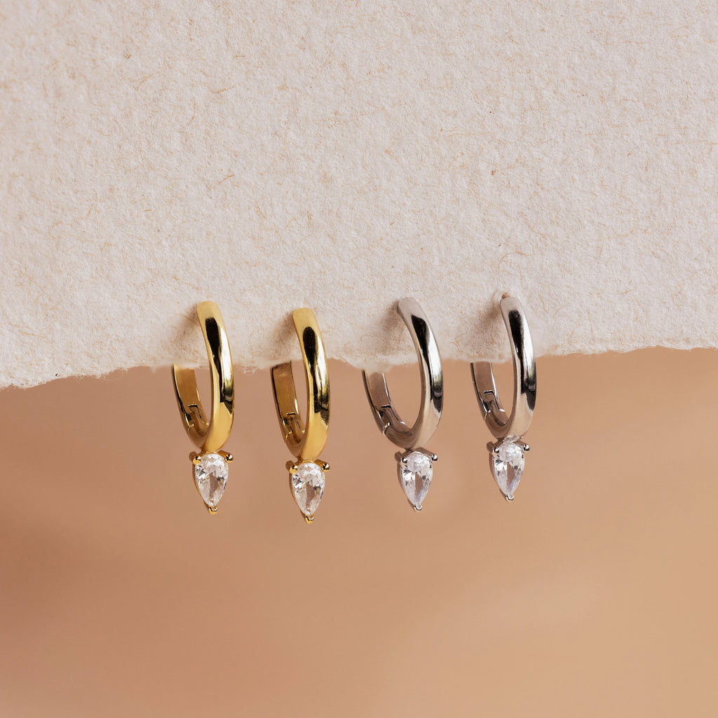 The Pear Diamond Drop Hoops set features two pairs of elegant hoops—one gold and one silver—each adorned with a clear teardrop-shaped gemstone, making them perfect for everyday elegance or as chic wedding accessories.